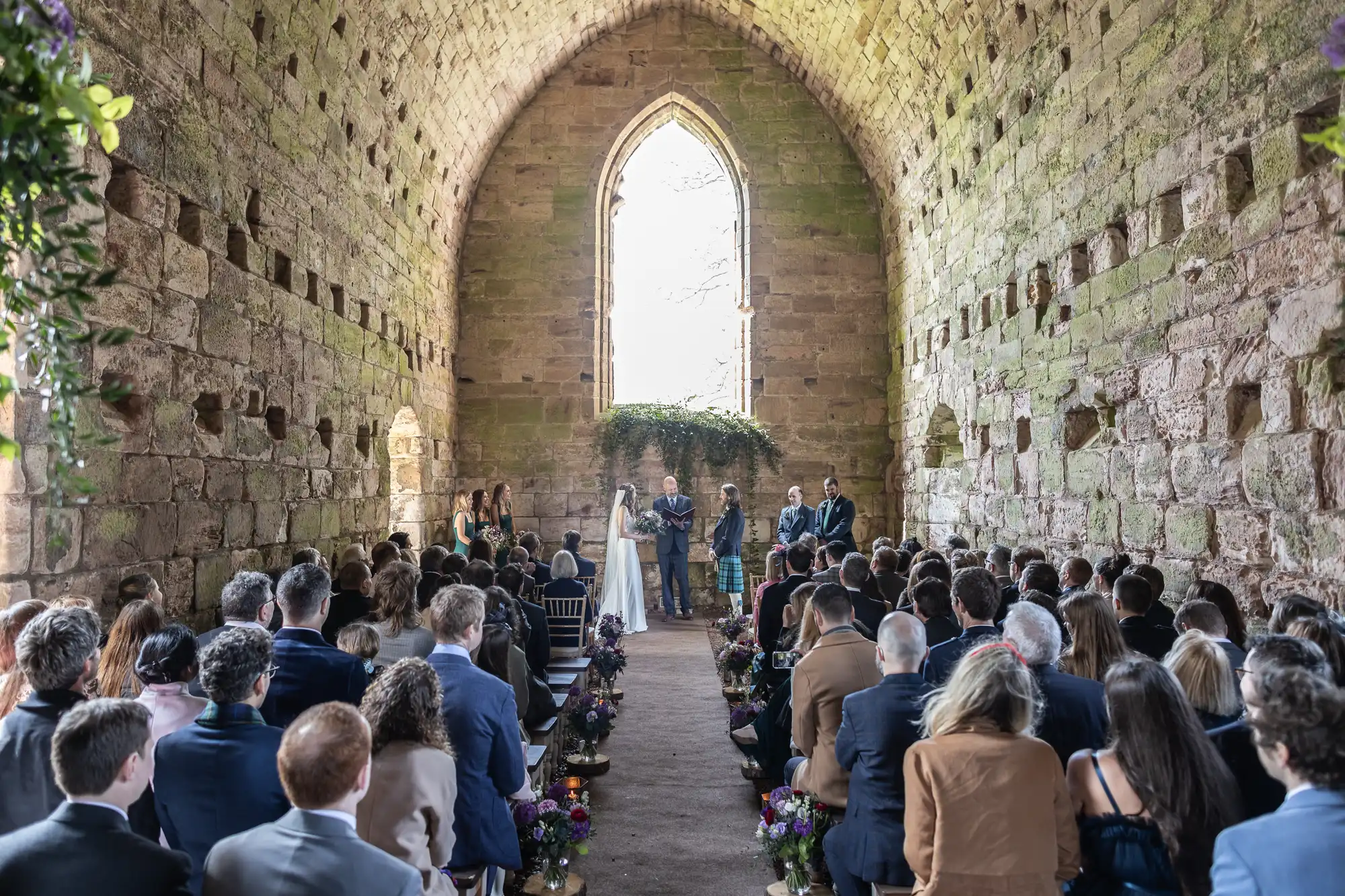 A wedding ceremony inside a stone chapel with high arched ceilings. Guests are seated on either side of a central aisle, and the couple stands before officiants near a large window.