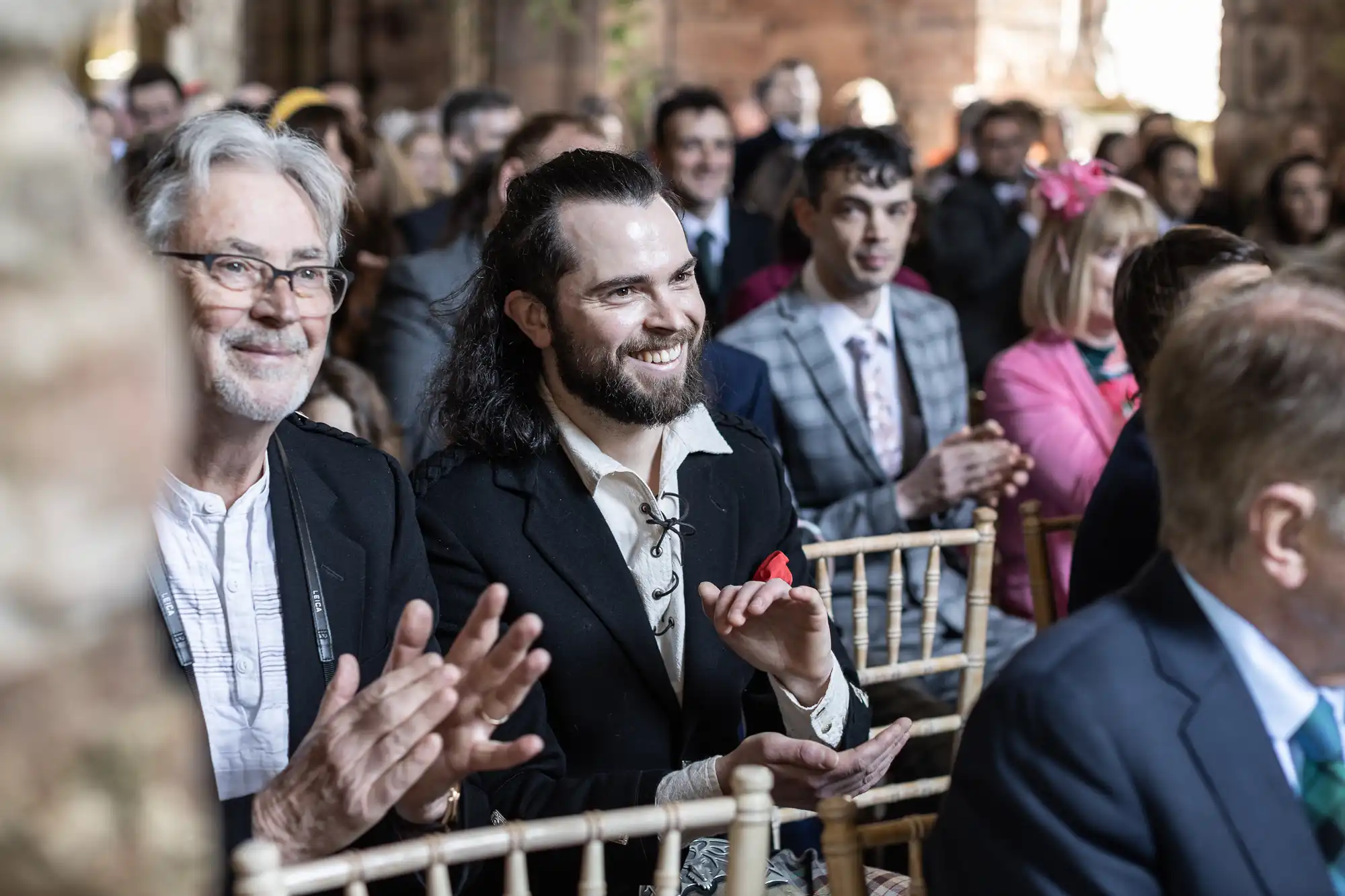 Group of people seated at an event, some clapping. The focus is on a smiling bearded man with long hair, wearing a black jacket and white shirt. The background shows other seated attendees.