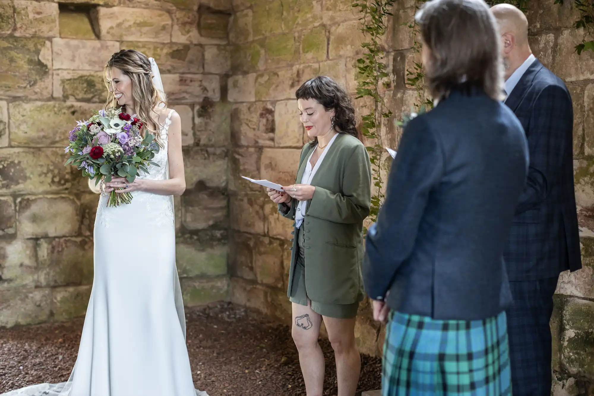 A woman in a wedding dress stands with a bouquet of flowers. Another woman reads from a paper while two people are in the foreground. The setting appears to be rustic with stone walls.