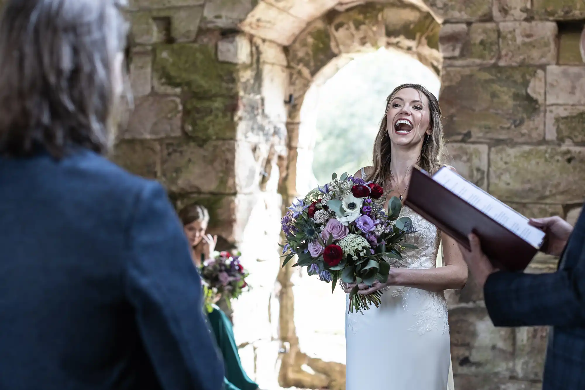 A bride holding a bouquet is laughing during a wedding ceremony in an old stone building with a large archway. Two other people are partially visible, one reading from a book.