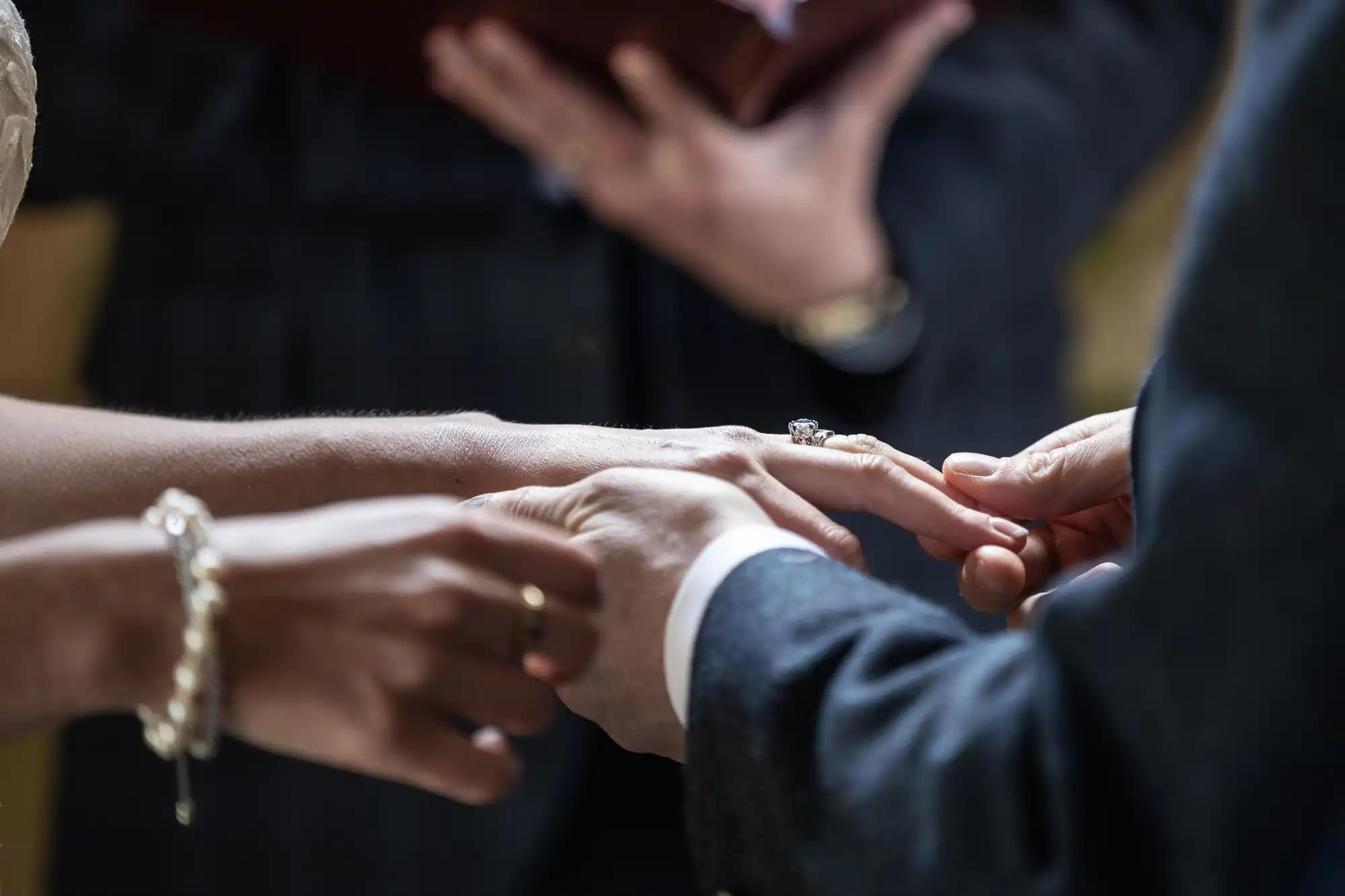Close-up of a person placing a ring on another person's finger during a ceremony, with the officiant holding a book in the background.
