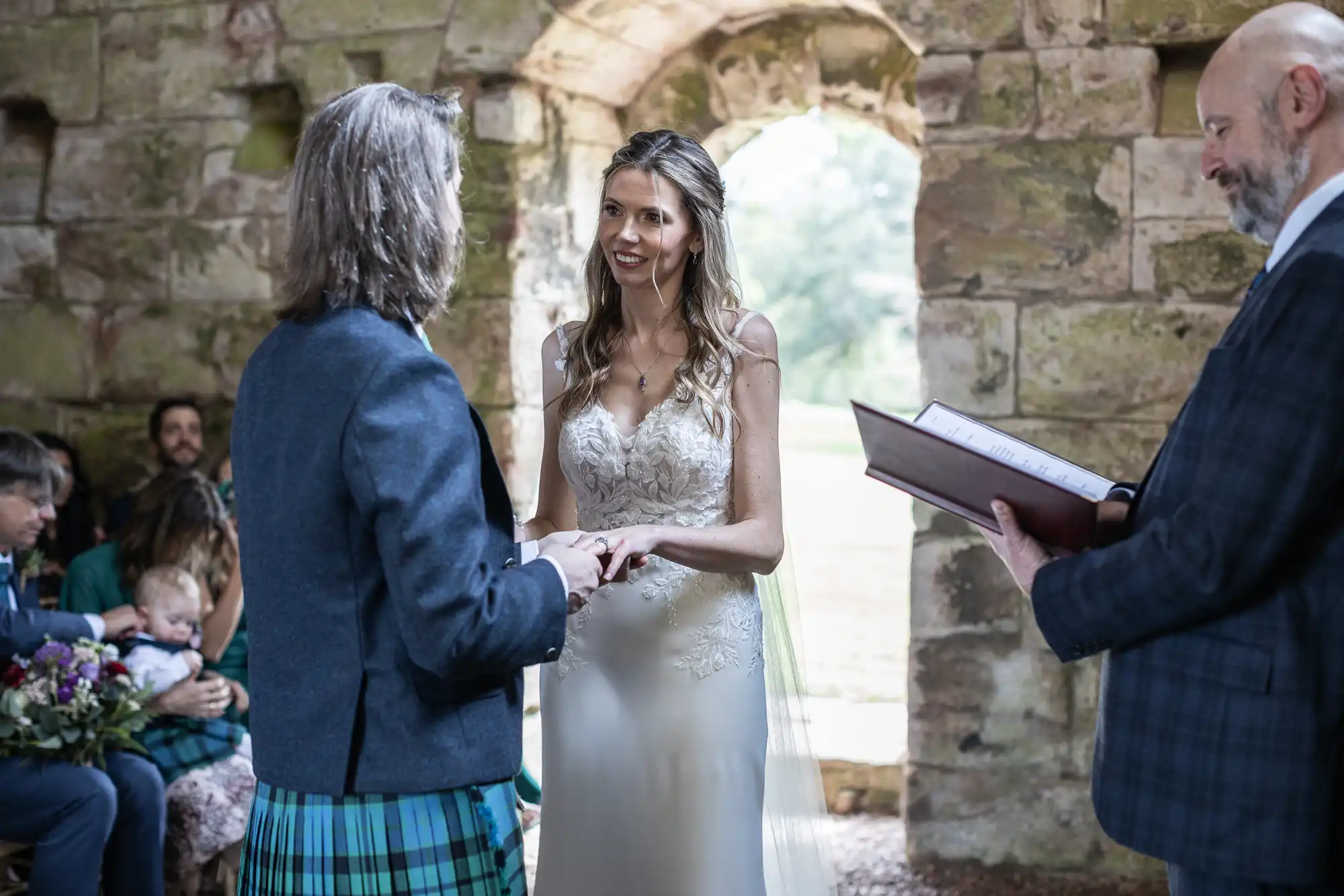 A couple stands in front of each other exchanging rings in a rustic stone building, while an officiant reads from a book. The bride wears a white gown and the groom wears a blue jacket with a kilt.