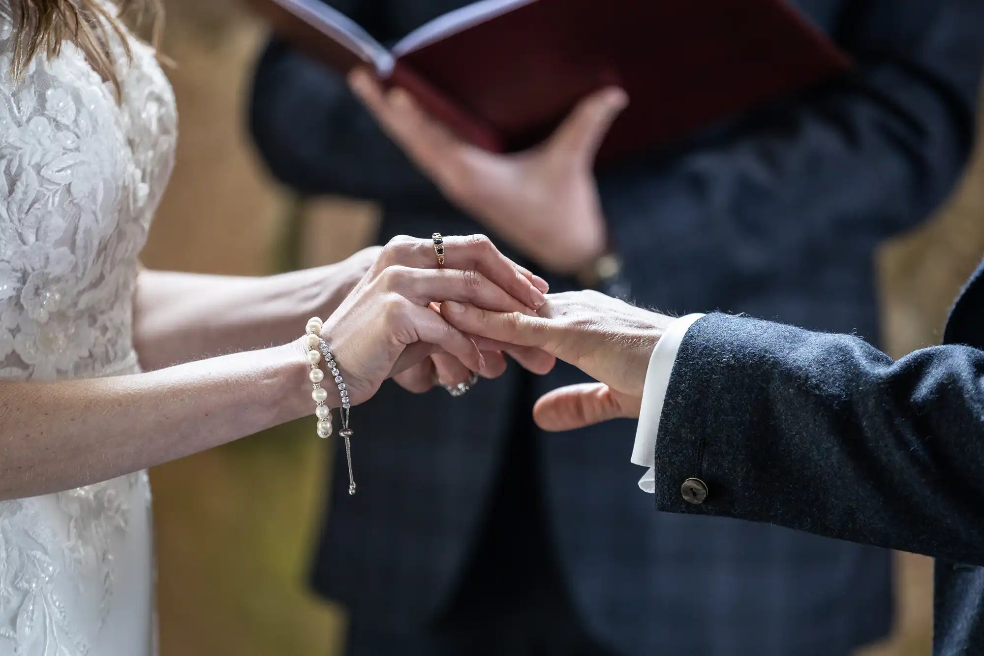 Close-up of a couple exchanging rings during a wedding ceremony, with an officiant holding an open book in the background. Both are dressed in formal attire.