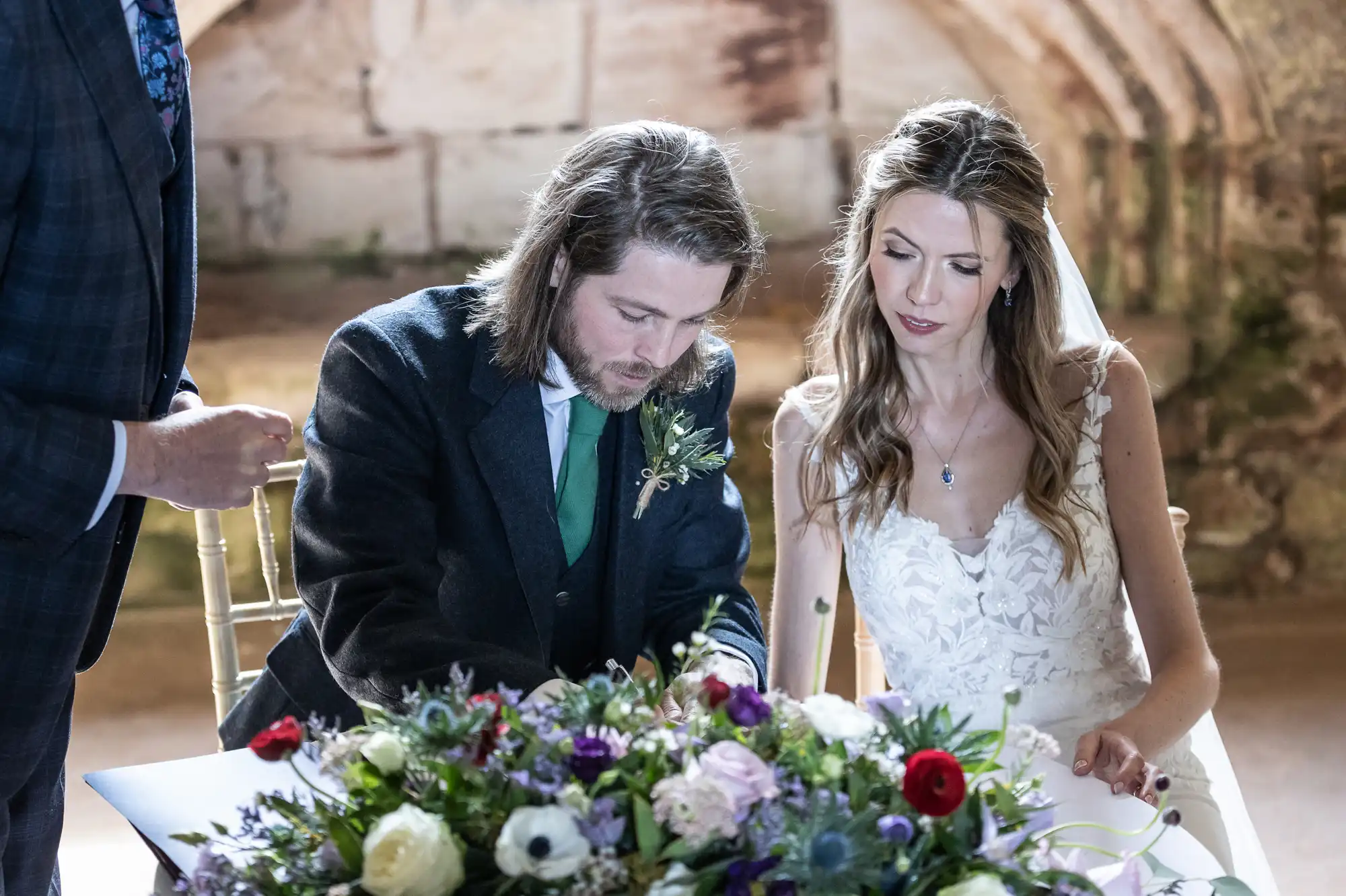 A couple is seated at a table, signing documents at their wedding. The groom wears a suit with a green tie and boutonniere, while the bride wears a white dress with lace details. A floral arrangement is on the table.