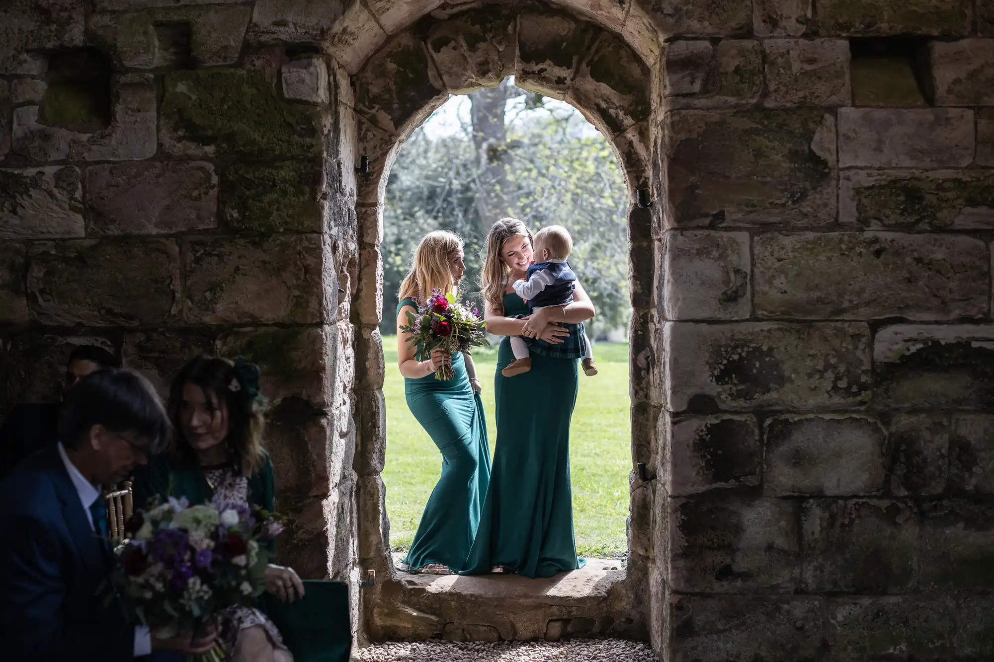 Two women in green dresses stand in a stone archway; one holds a bouquet of flowers and the other holds a child. People are seated with flowers in the foreground.