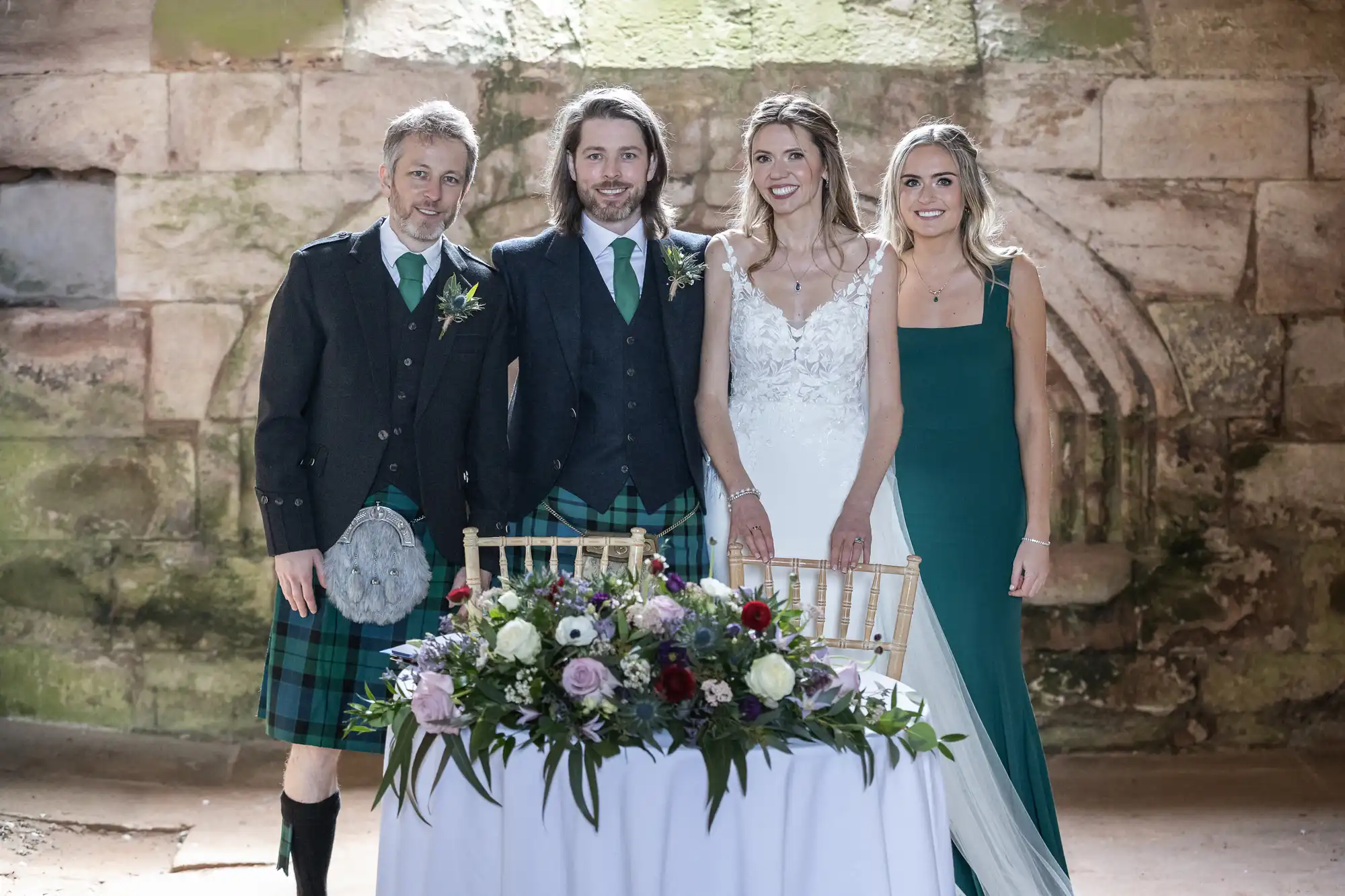Four people, two men in kilts and two women in dresses, stand behind a table with a floral arrangement in what appears to be a stone building.