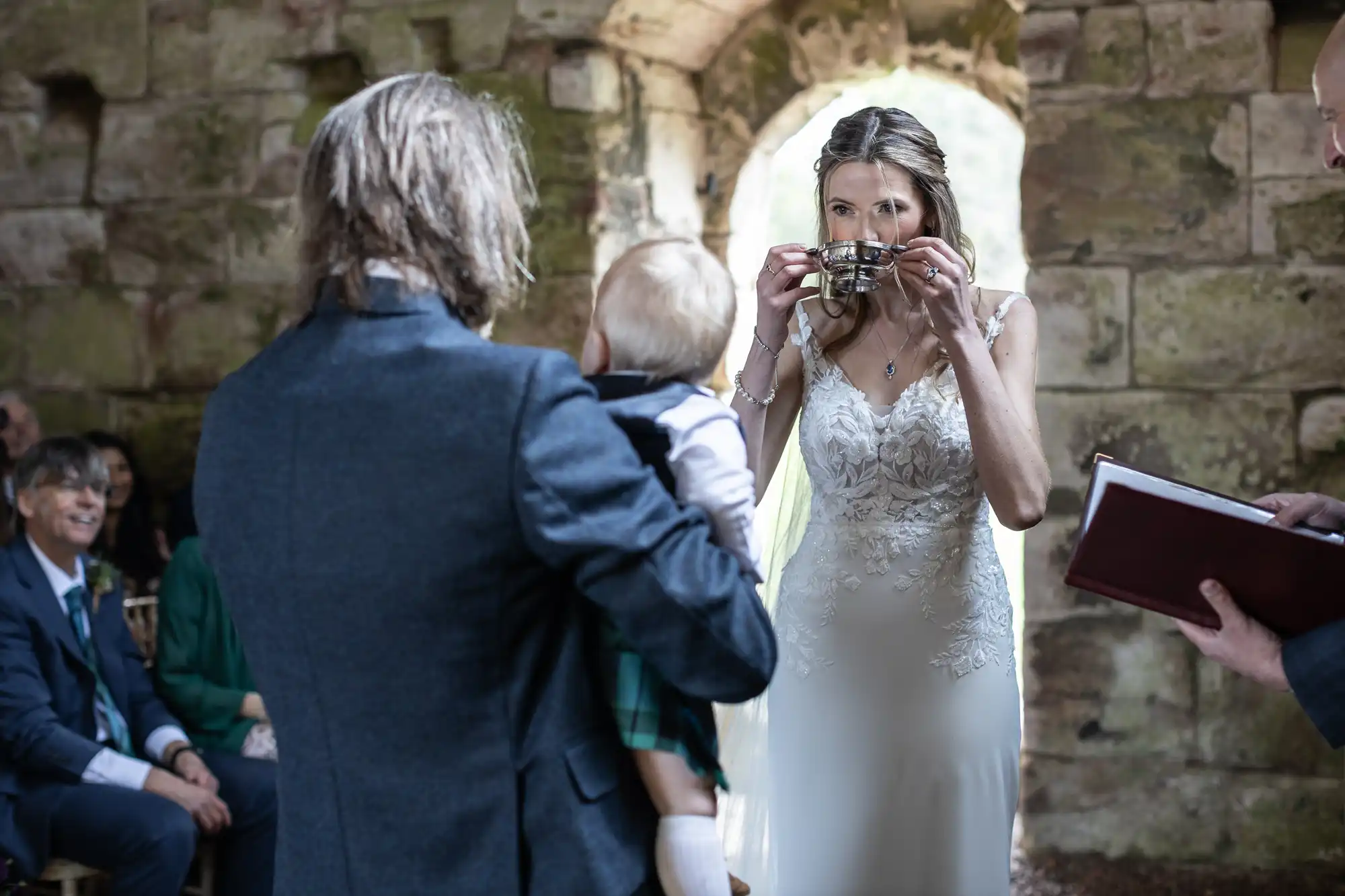 Bride in a white dress drinks from a goblet, while a groom in a blue suit holds a baby, in a stone-walled setting, with guests watching and an officiant holding a book.