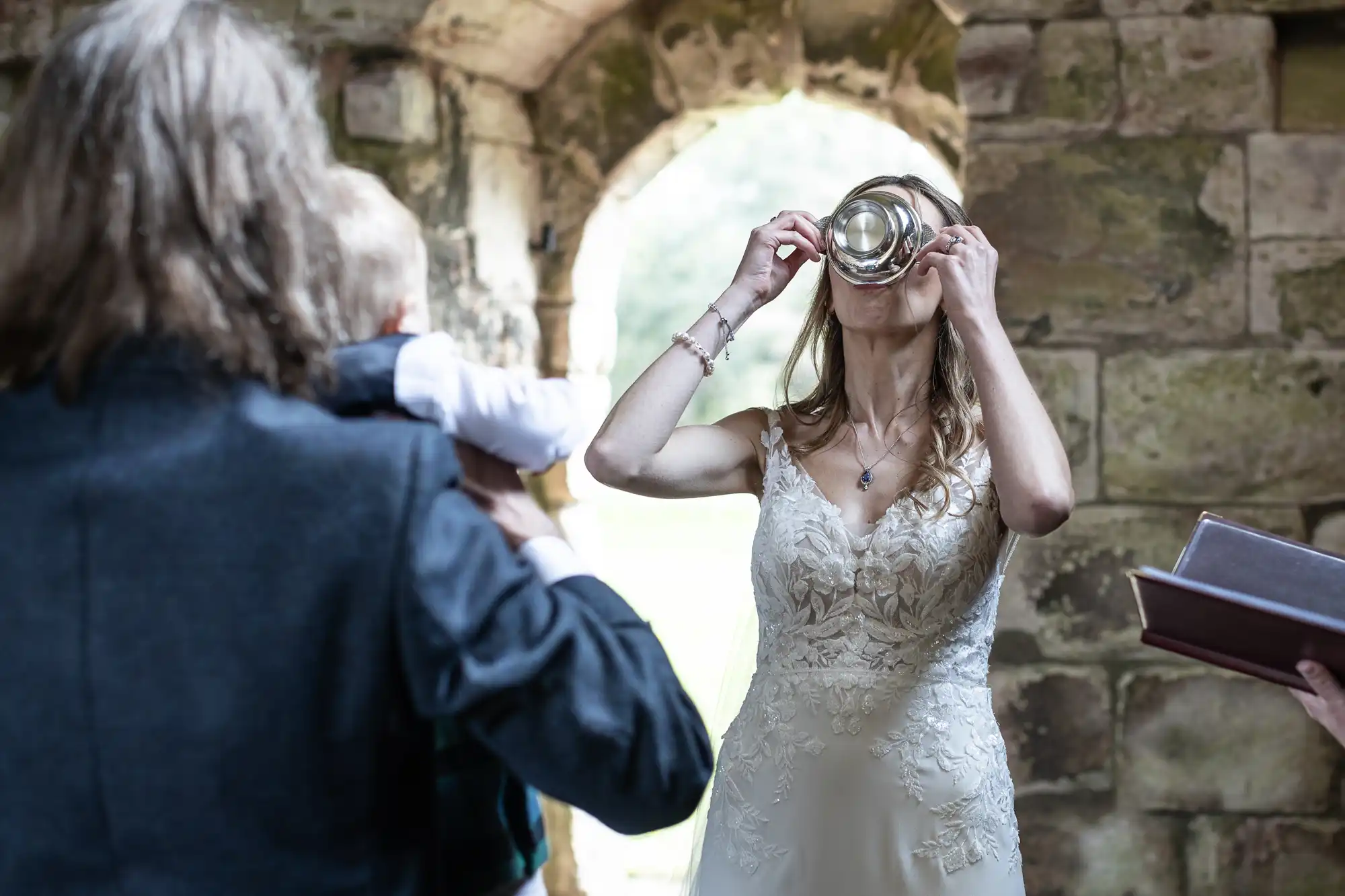 A bride in a wedding dress drinks from a metal cup, while a man holding a child looks on in what appears to be a rustic stone building.