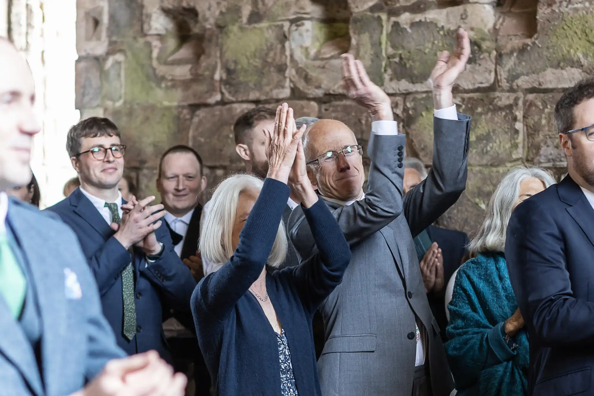 A group of people in formal attire stands inside a stone building, clapping and raising their hands in applause.