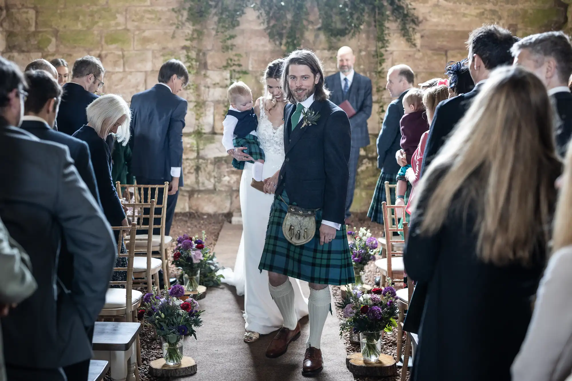 A couple walks down the aisle in a wedding ceremony. The groom wears a kilt, while the bride holds a child. Guests line the aisle, and a stone wall with foliage is in the background.