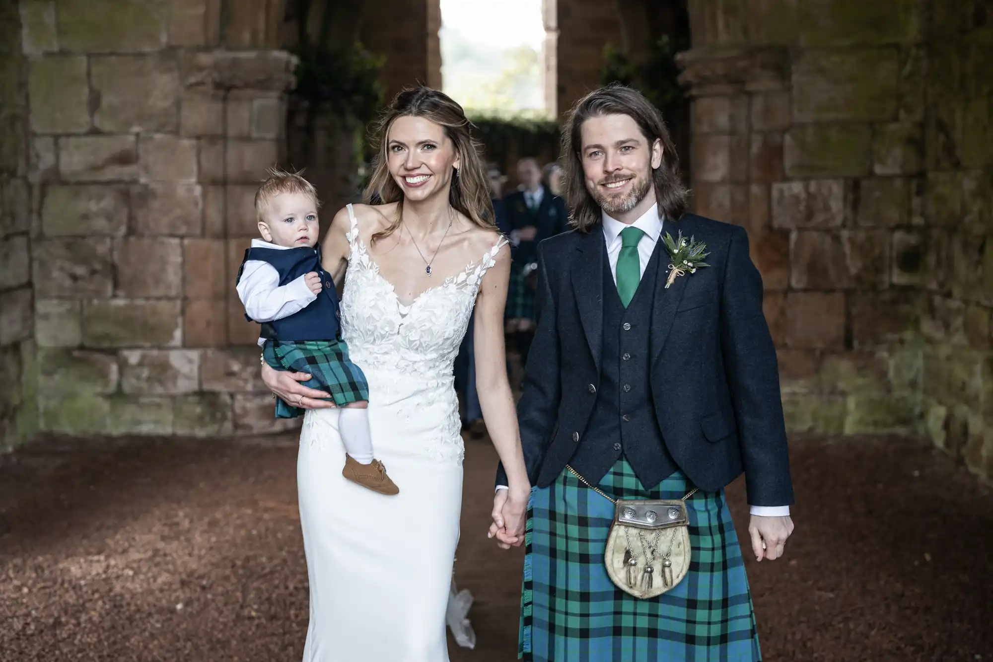A couple dressed in wedding attire, with the man and child wearing traditional Scottish kilts, stand together holding hands in front of a rustic stone structure. The woman holds the child in her left arm.