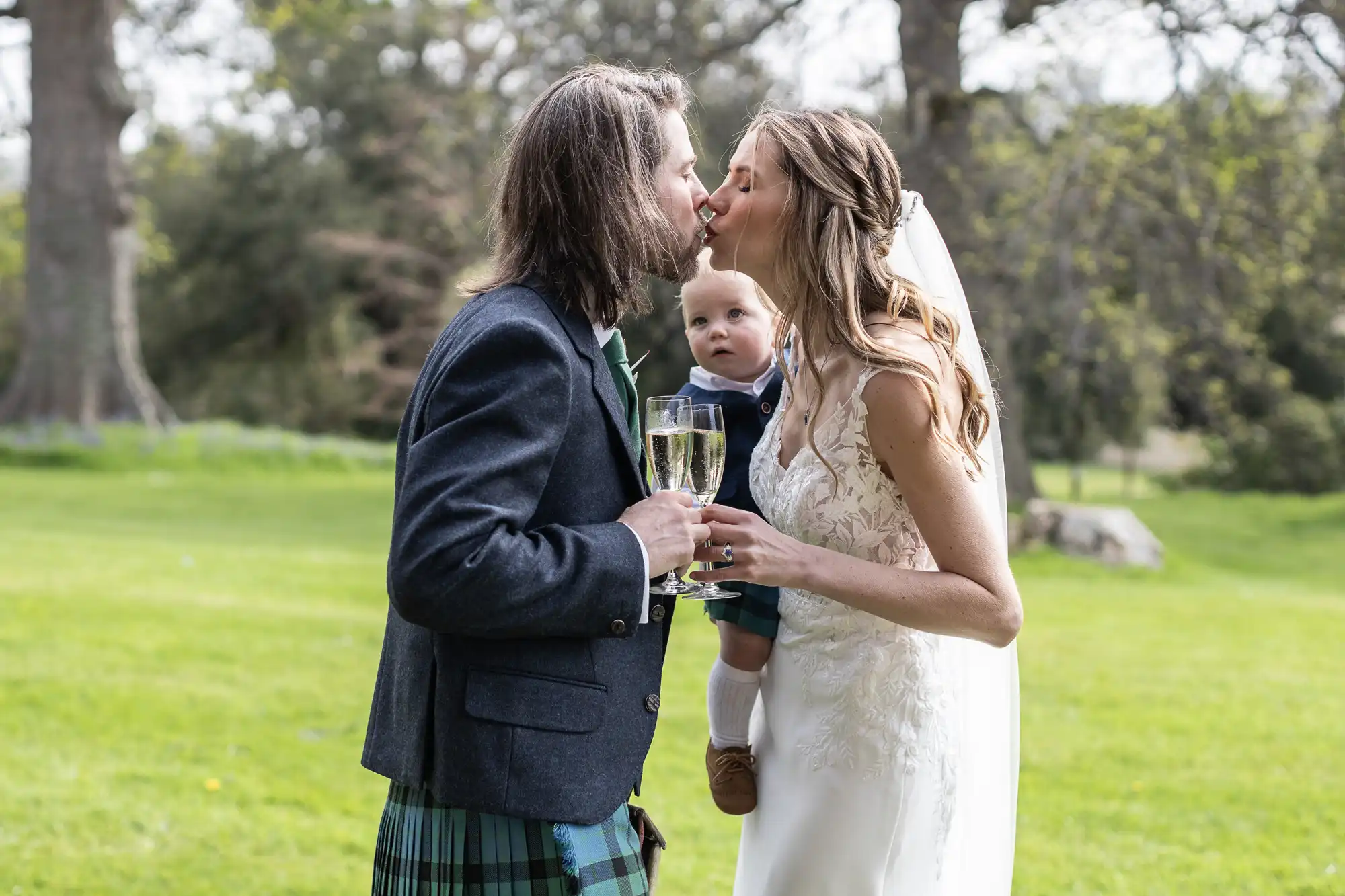 A couple in wedding attire kiss while holding champagne glasses, with the bride holding a baby between them. They stand on a grassy area with trees in the background.