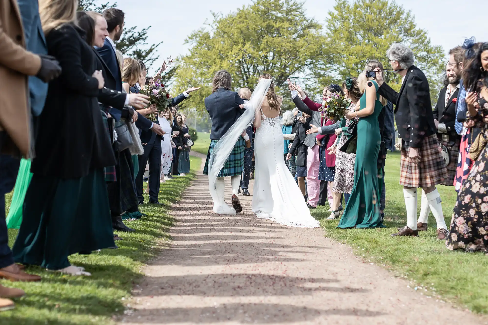 A newlywed couple walks down a path while guests, standing on both sides, applaud and take photos. The bride is in a white dress, and the groom is in a traditional kilt. Trees are in the background.