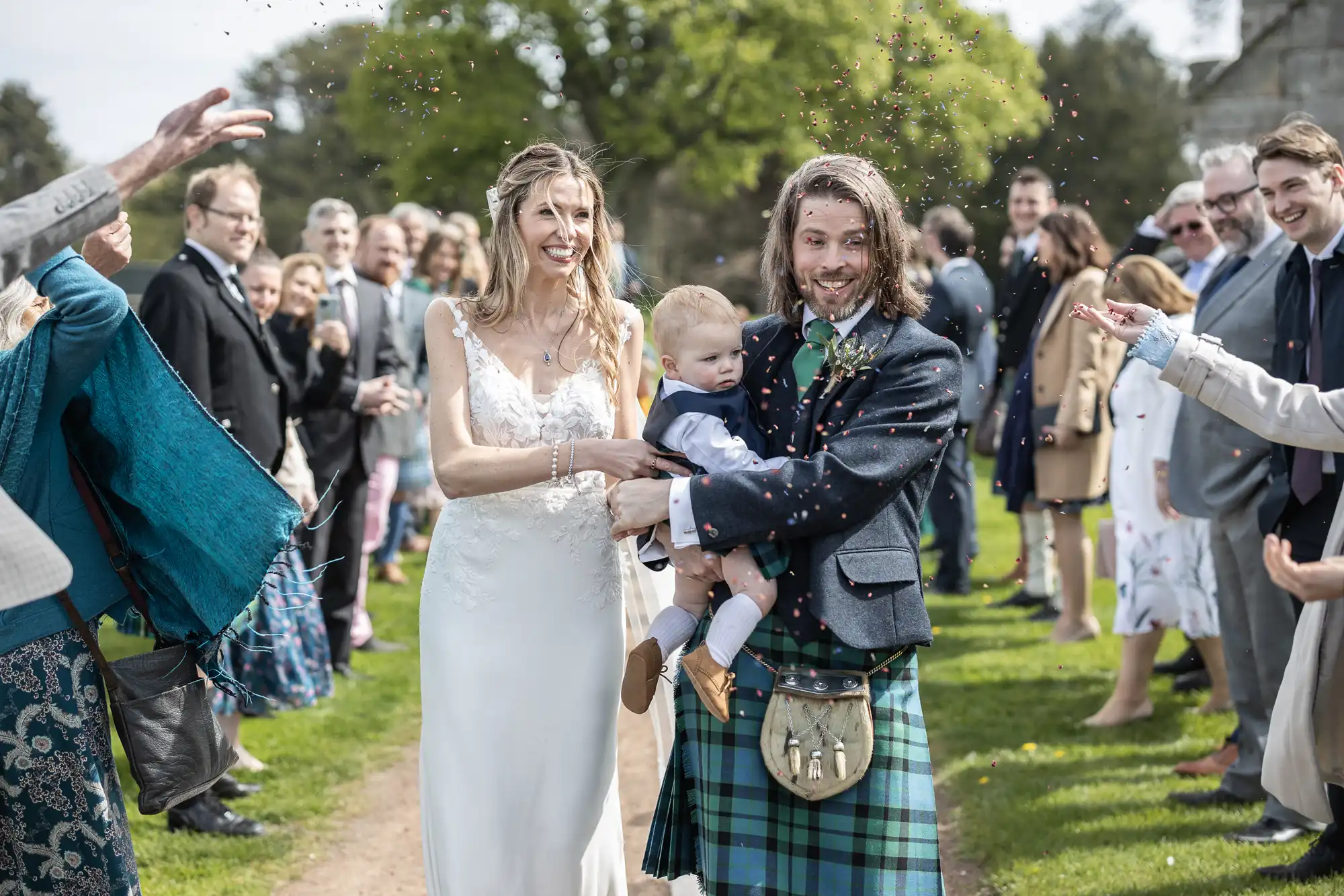 A bride and groom, holding a baby, walk down an outdoor aisle while guests celebrate by throwing confetti. The groom is wearing a kilt, and the bride is in a white gown.