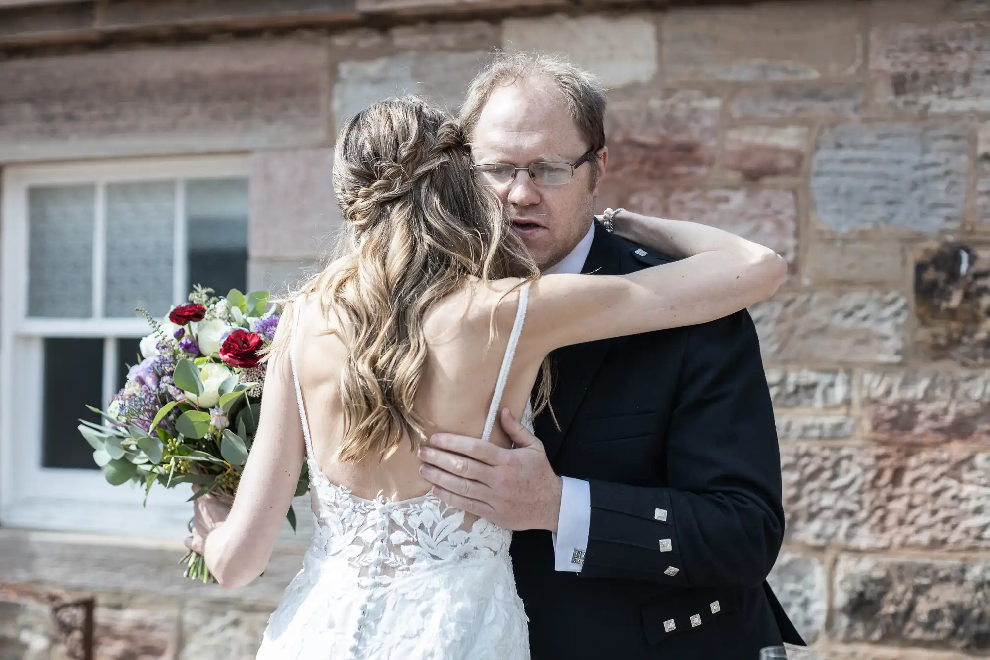 A bride in a white lace dress hugs a man in a formal black suit while holding a bouquet of flowers. They are outside in front of a brick building.