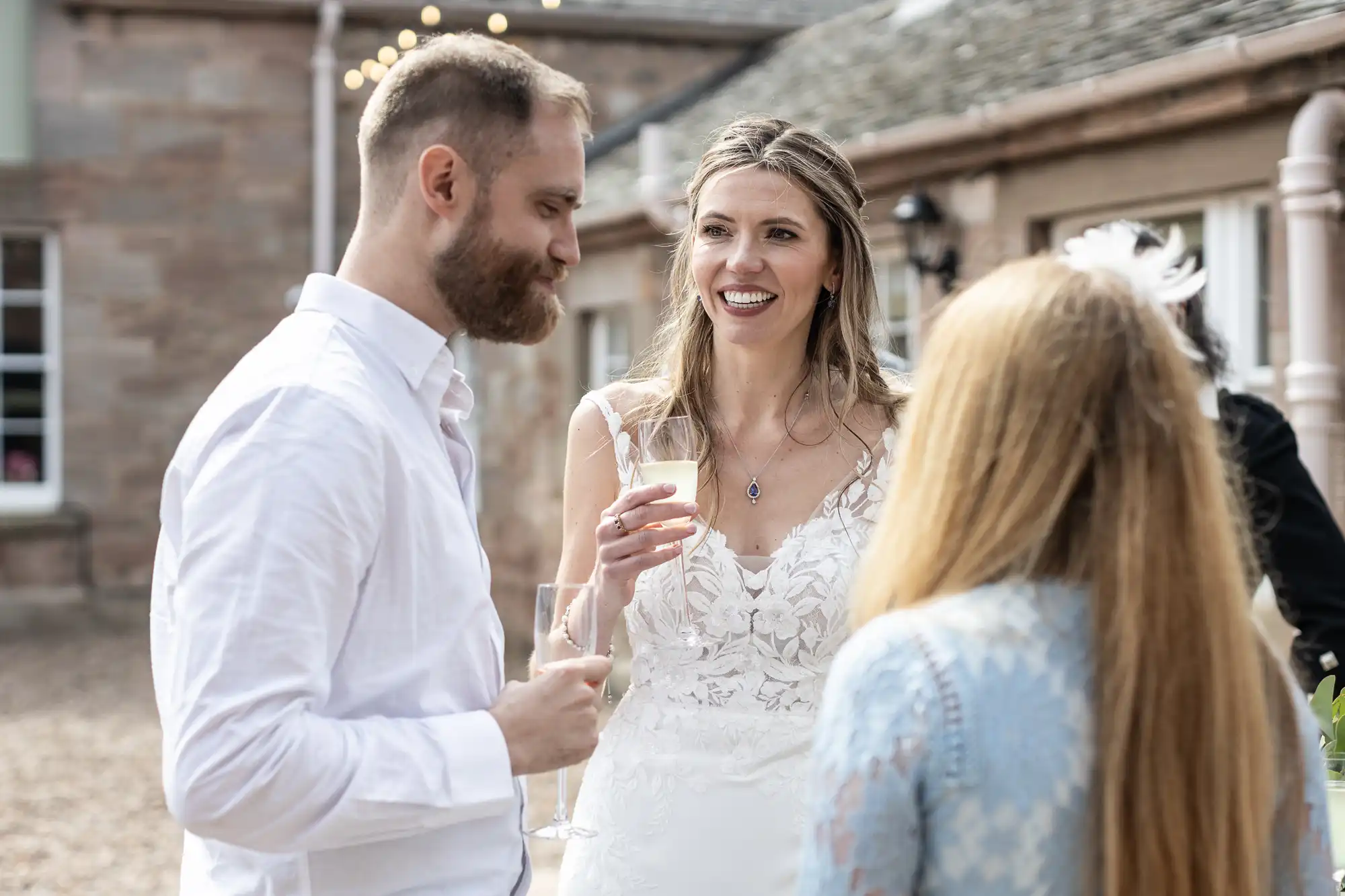 A bride in a white dress holds a glass and smiles while talking to a bearded man and a woman with long hair at an outdoor event.