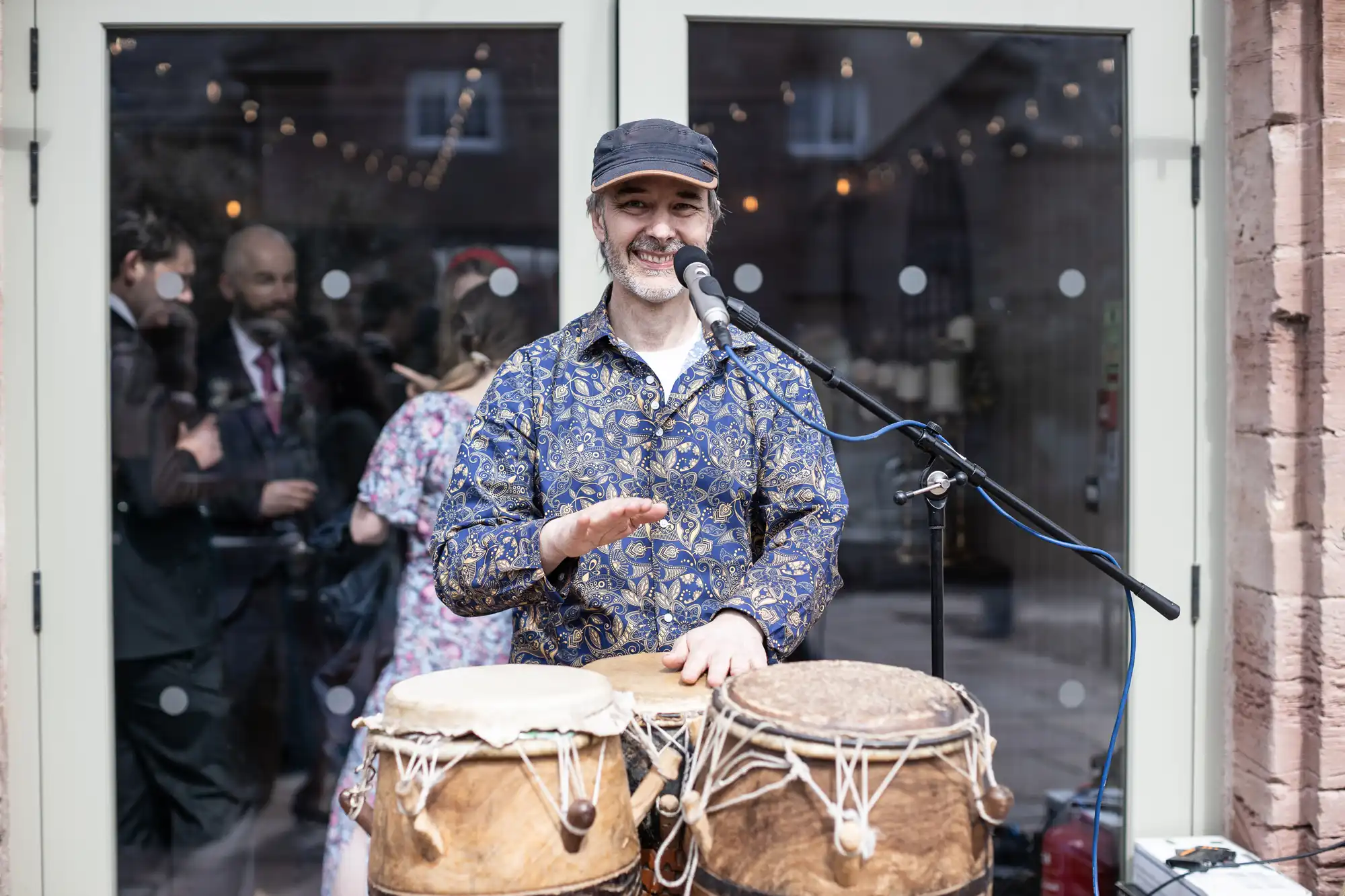 A man wearing a patterned shirt and cap is playing conga drums and smiling at a microphone in front of a glass door. People are conversing in the background.