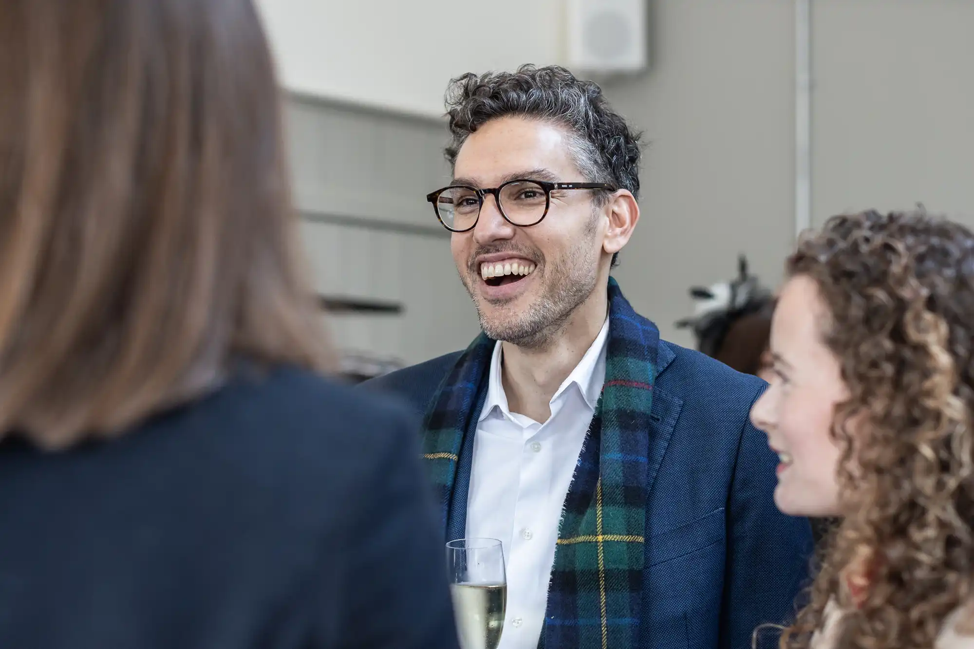 A man with glasses and a plaid scarf smiles while holding a drink, engaging in conversation with two other people.