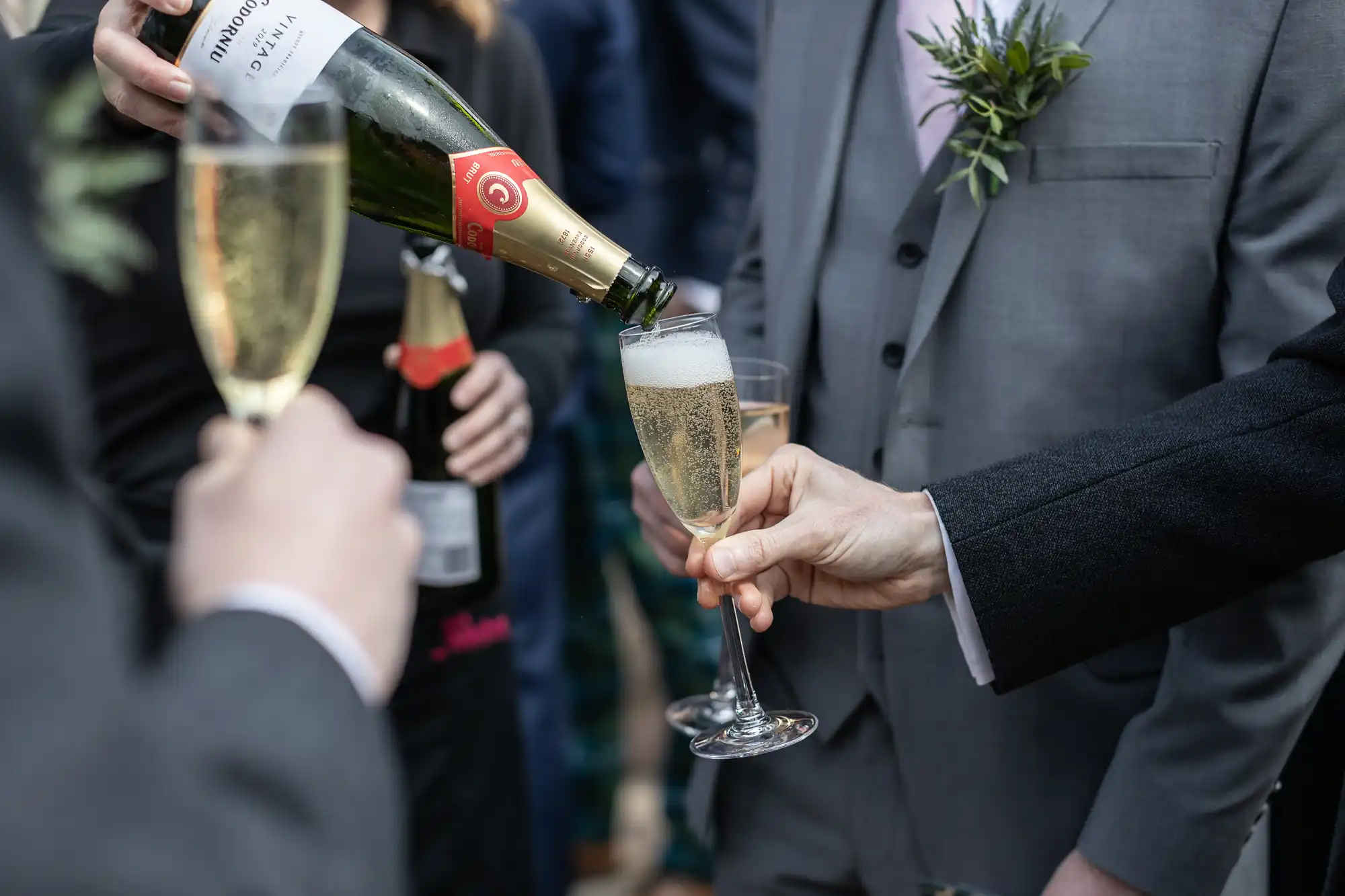 Hands holding champagne flutes as champagne is being poured, with people in formal attire in the background.