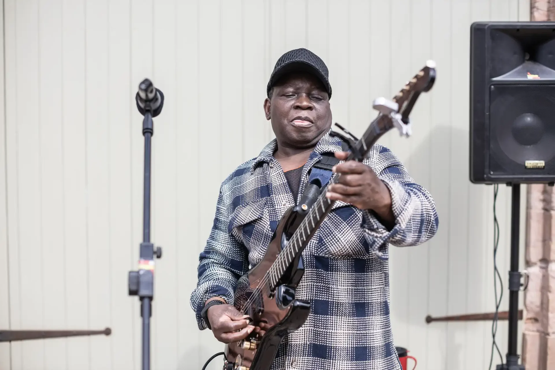 A man in a checkered shirt and cap plays an electric guitar on stage next to a microphone stand and speaker.