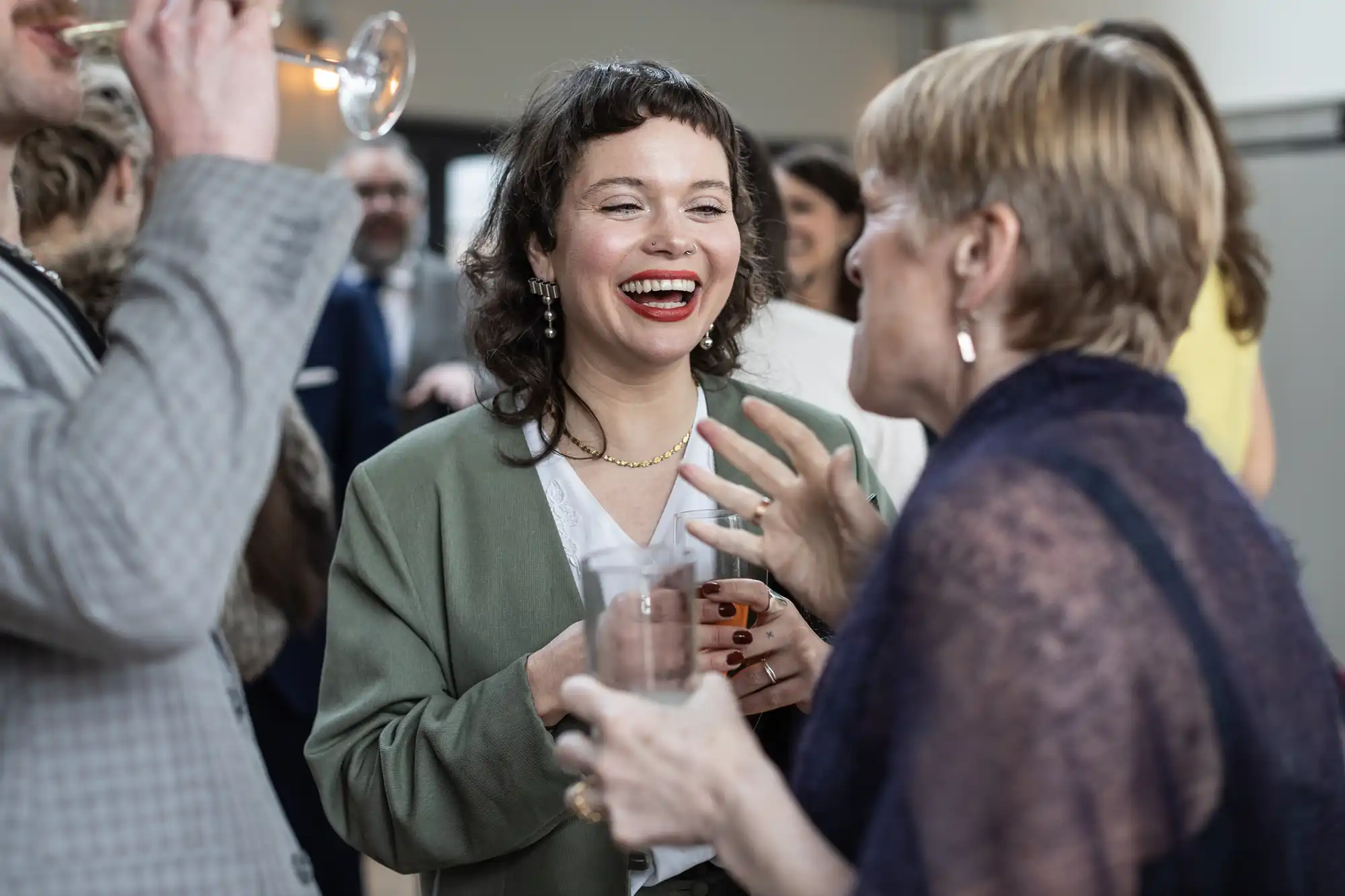 Two women engage in a lively conversation at a social gathering. One holds a drink and smiles broadly, while the other gestures expressively. Other guests are visible in the background.