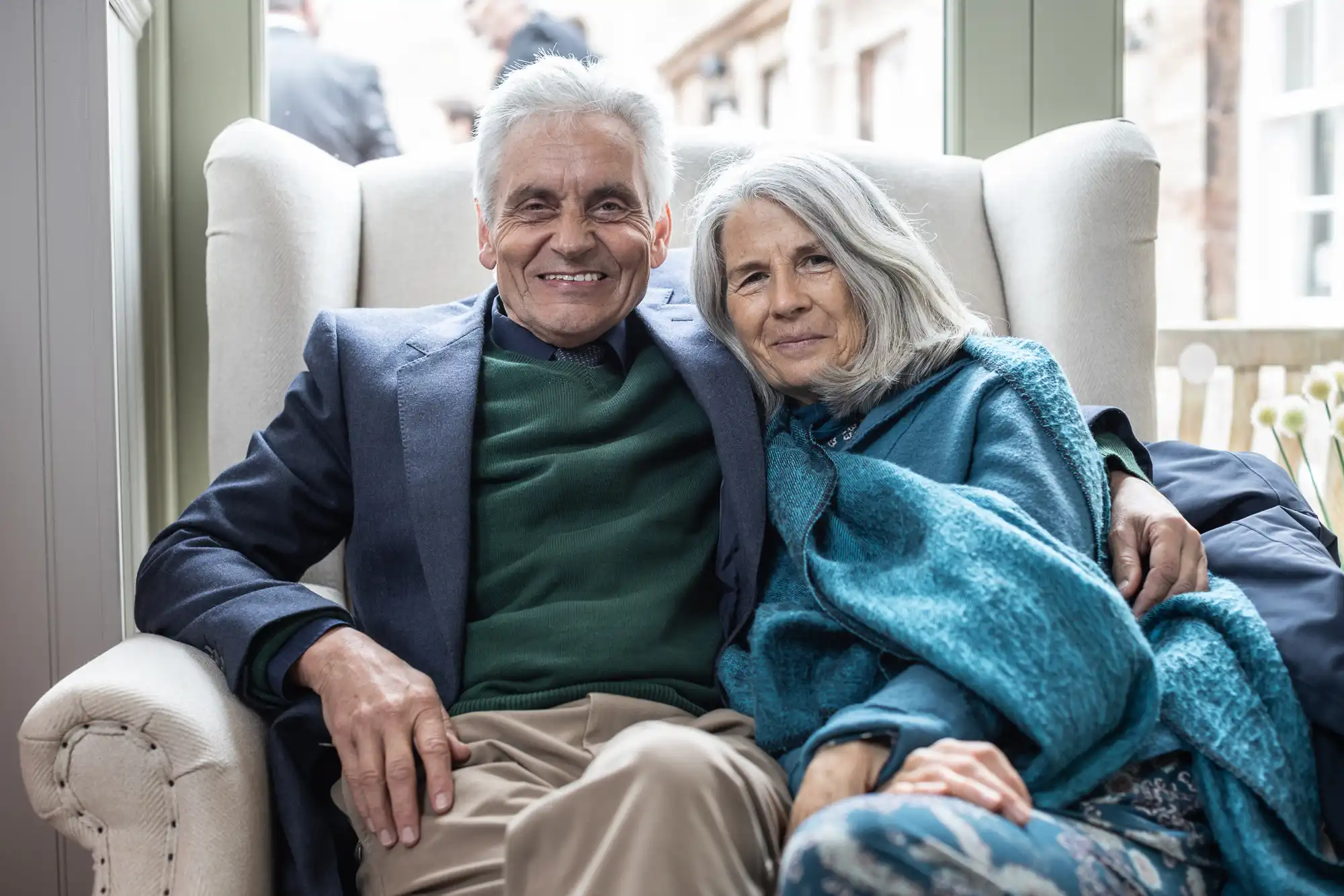 An elderly man and woman sit closely together on an armchair, smiling warmly at the camera. The woman is wrapped in a blue blanket, and they appear to be in a well-lit indoor setting.