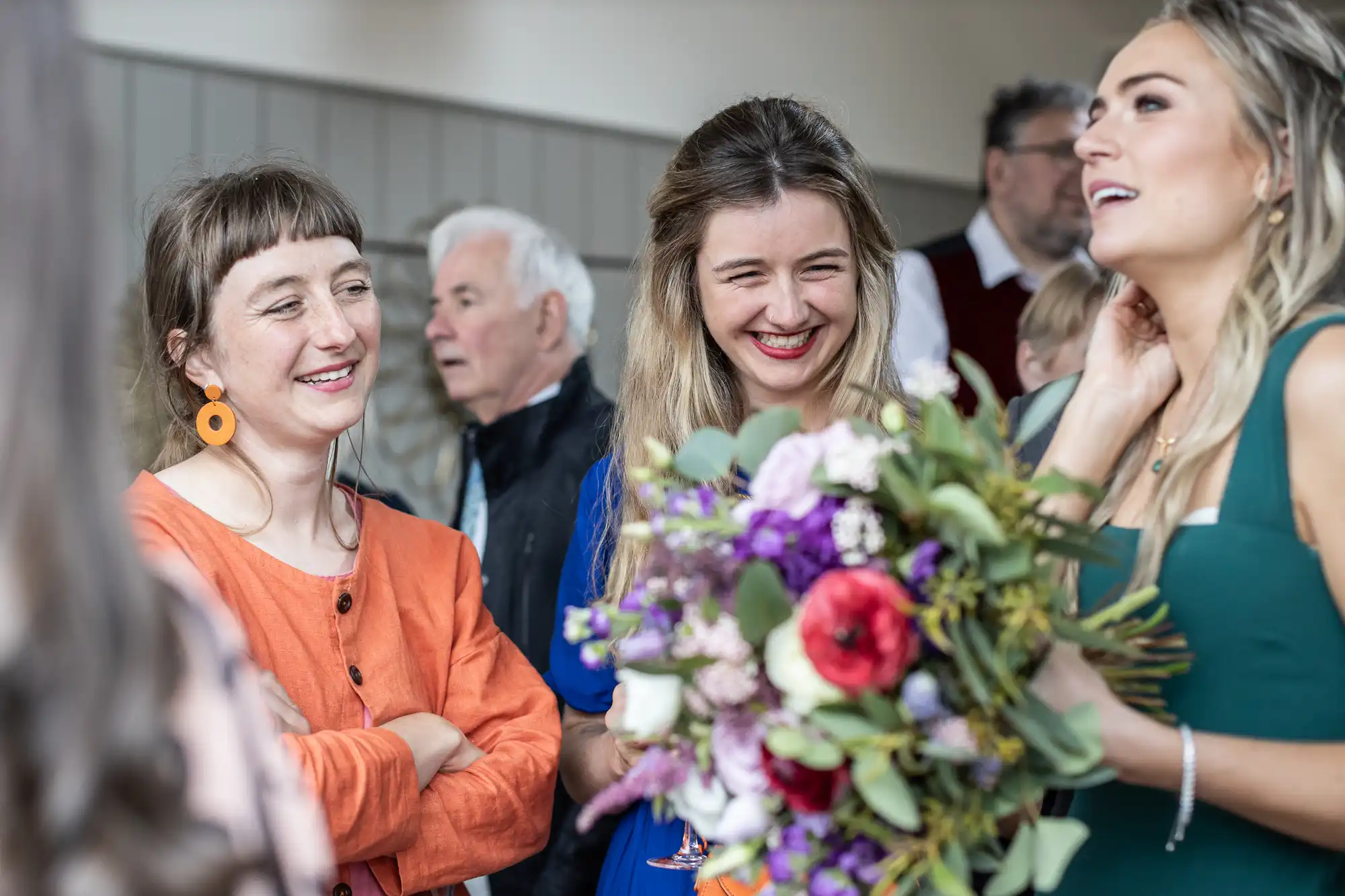 Three women, one holding a bouquet, are smiling and talking at an indoor gathering. Several other people are visible in the background.