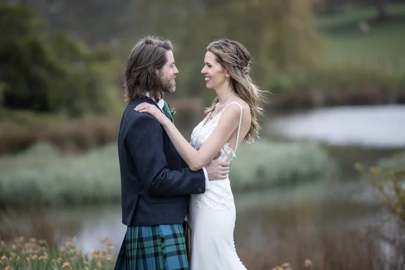 wedding photography at Dunglass estate - A couple is standing outdoors, facing each other and smiling. The man is wearing a kilt and suit jacket, while the woman is wearing a white dress. A body of water and greenery are in the background.
