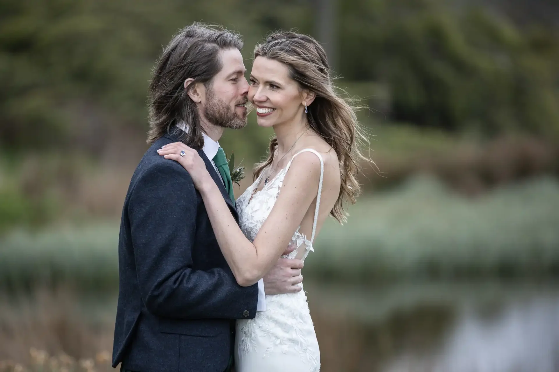 A couple embracing outdoors, the man in a dark suit and the woman in a white sleeveless dress, with a natural background of greenery and water.