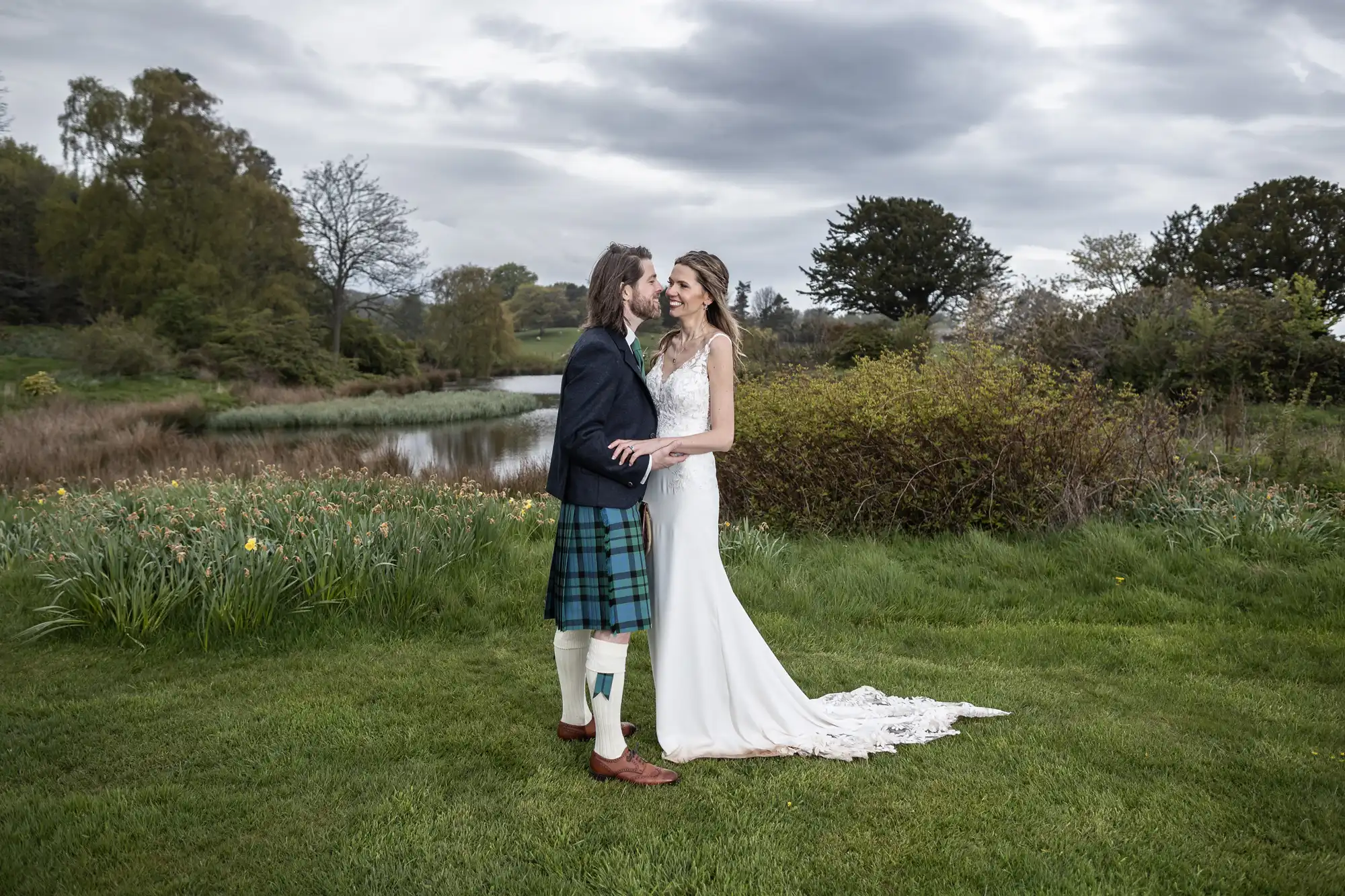 A couple dressed in wedding attire stands on a grassy area near a pond, with the man wearing a kilt and the woman in a white gown, under a cloudy sky.