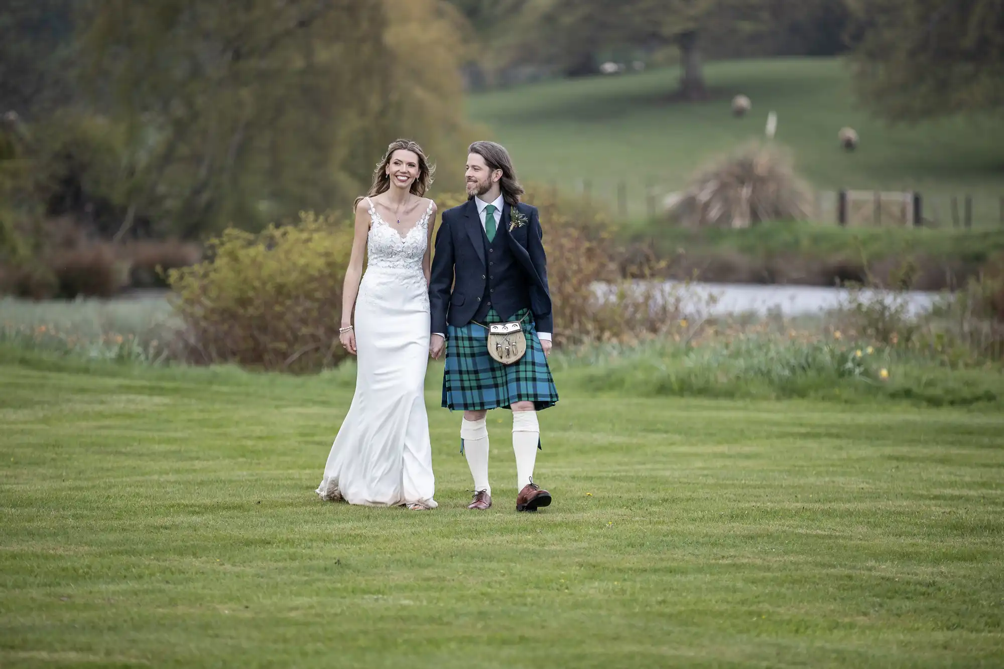A bride in a white dress and groom in a kilt walk hand in hand on a grassy field with lush greenery and a body of water in the background.