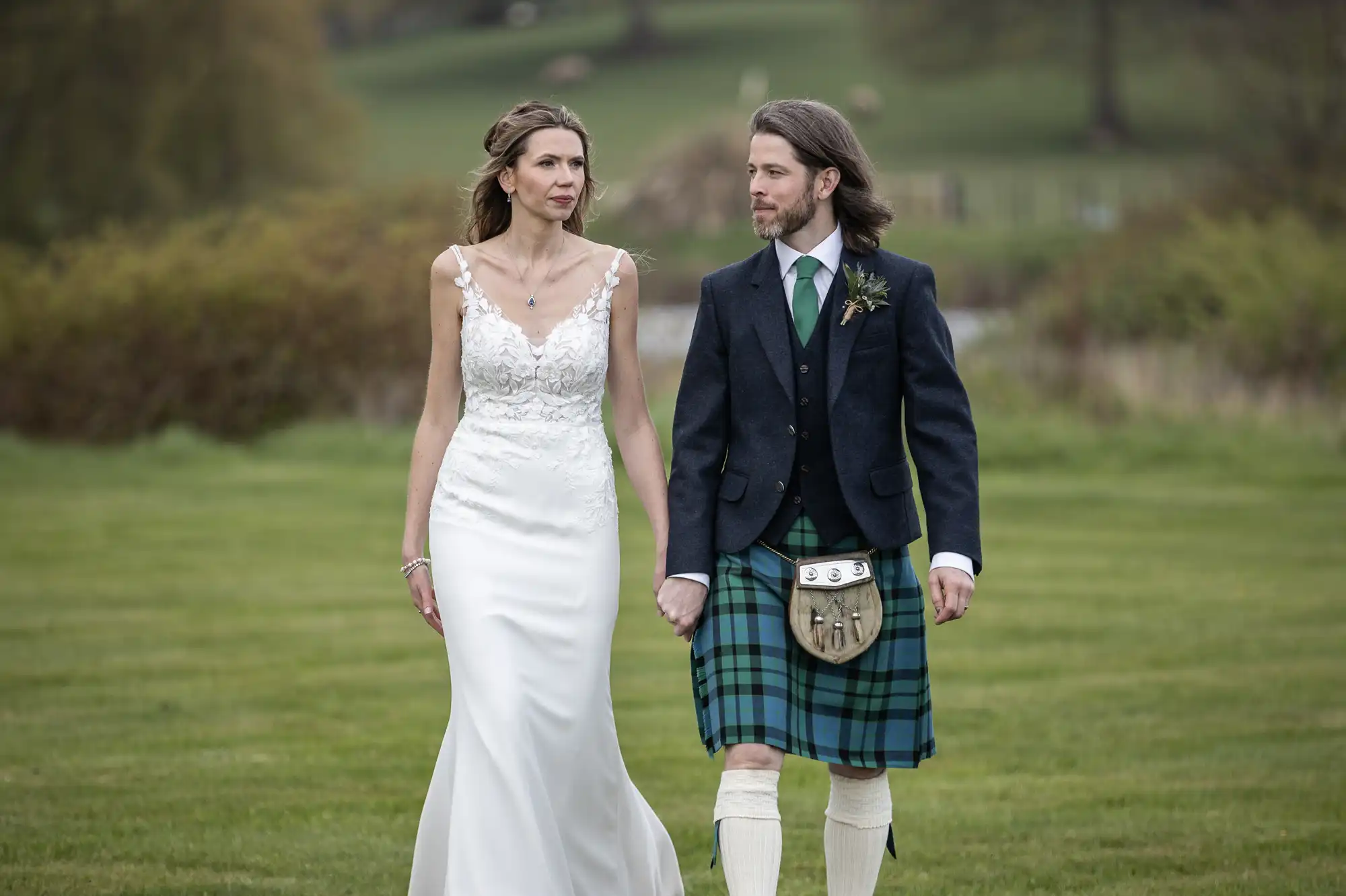A bride in a white dress and a groom in a kilt walk hand-in-hand on a grassy field.