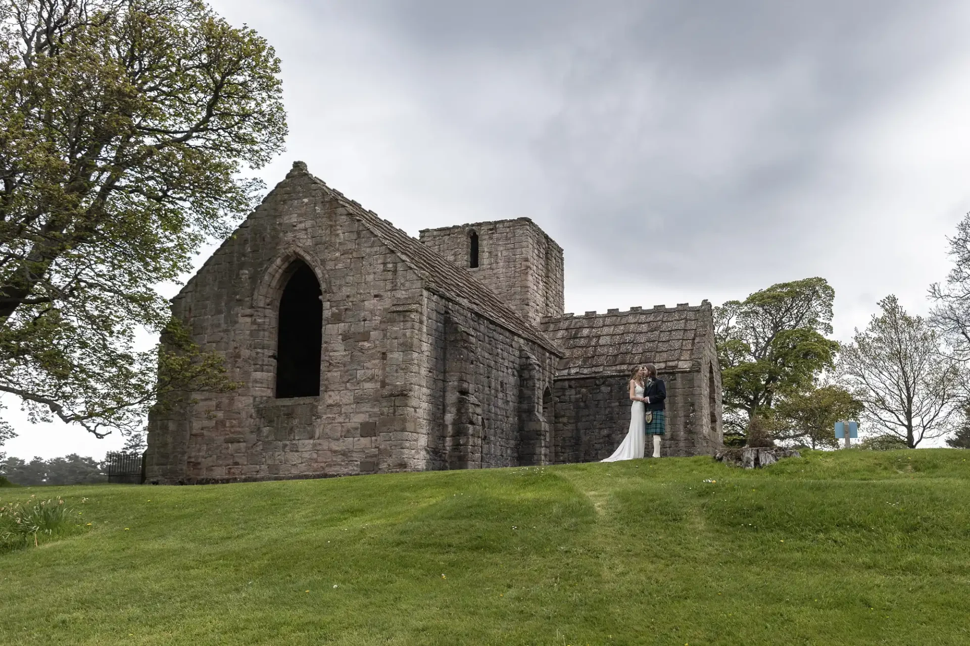 A couple stands in front of a historic stone building with a large arched window, surrounded by green grass and trees under a cloudy sky.