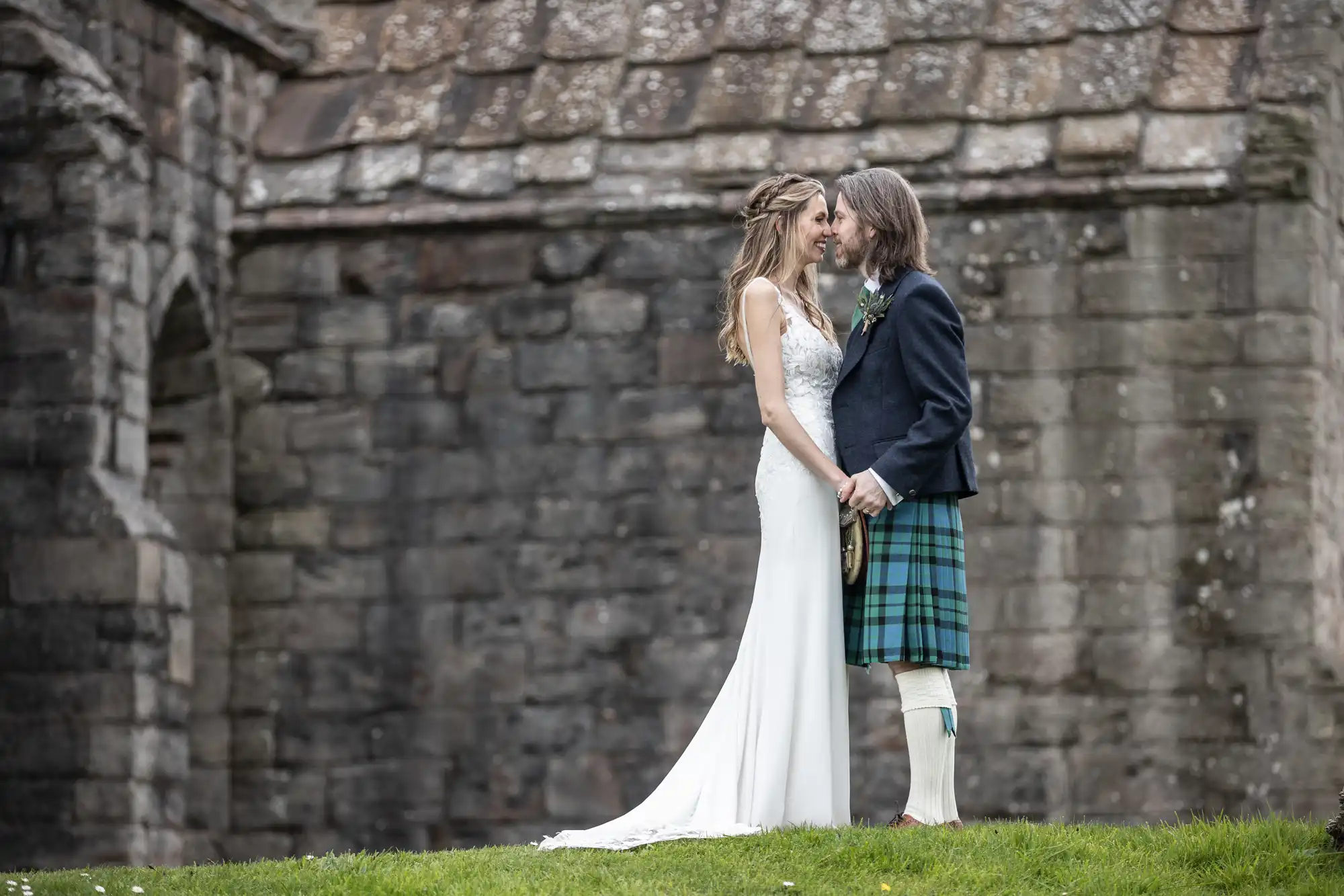 A couple stands holding hands and looking at each other in front of a stone building. The woman is in a white dress, and the man is in a traditional Scottish kilt.