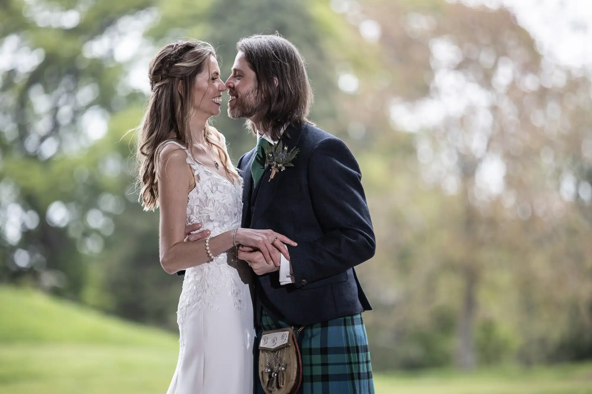 A bride and groom stand close, holding hands and smiling at each other. The groom wears a kilt, and the bride is in a white wedding dress. They are outdoors with greenery in the background.