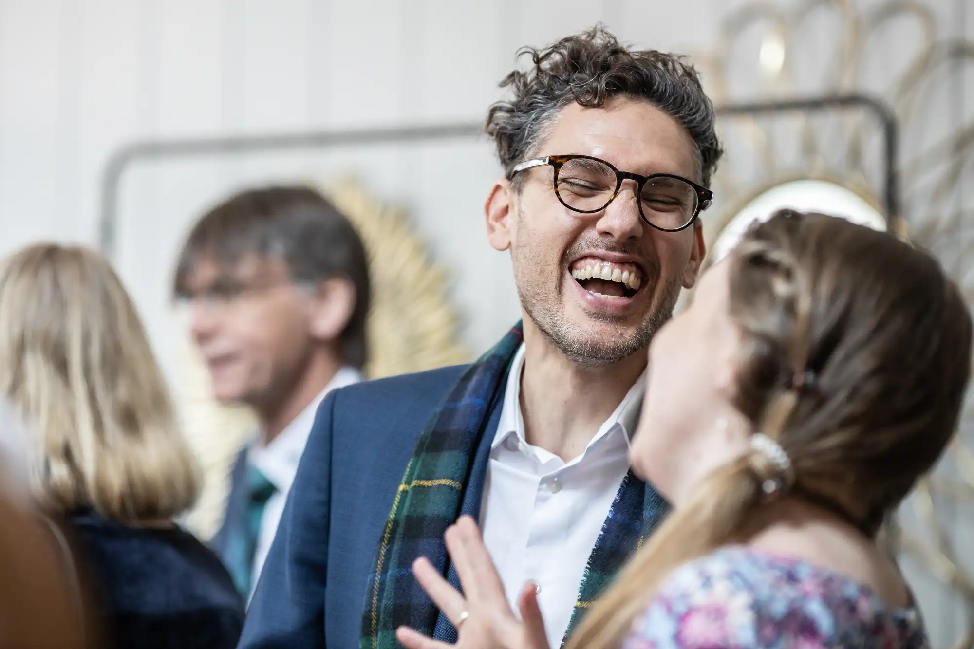 A man with glasses and curly hair laughs with a woman at a social gathering. Other people are visible in the background.