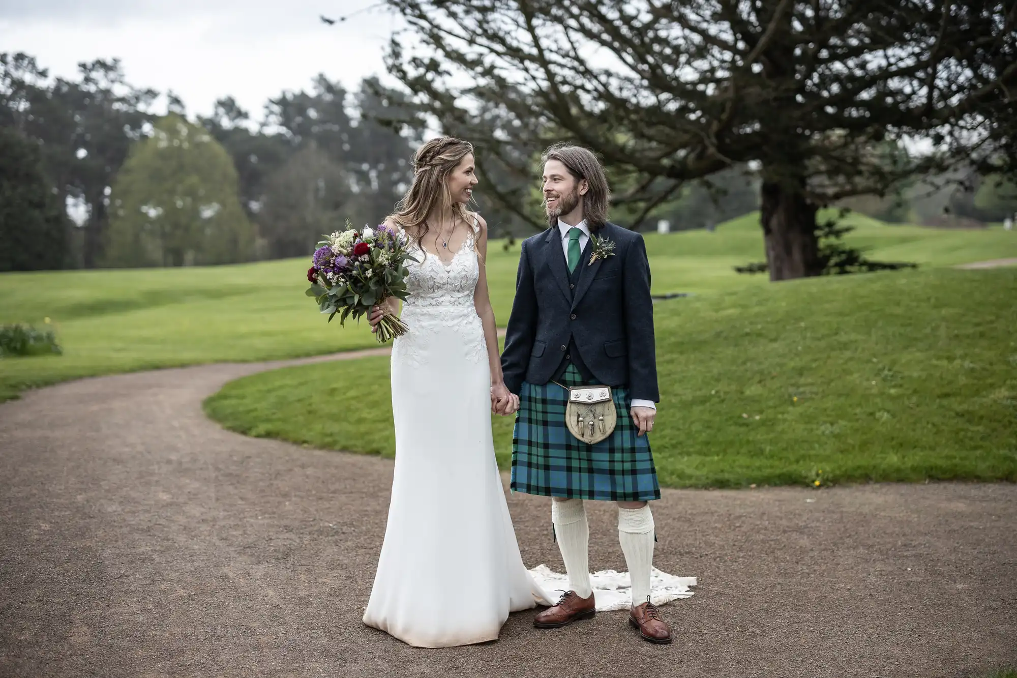 A bride in a white dress and a groom dressed in traditional Scottish attire hold hands and smile at each other, standing on a path in a green, park-like setting.