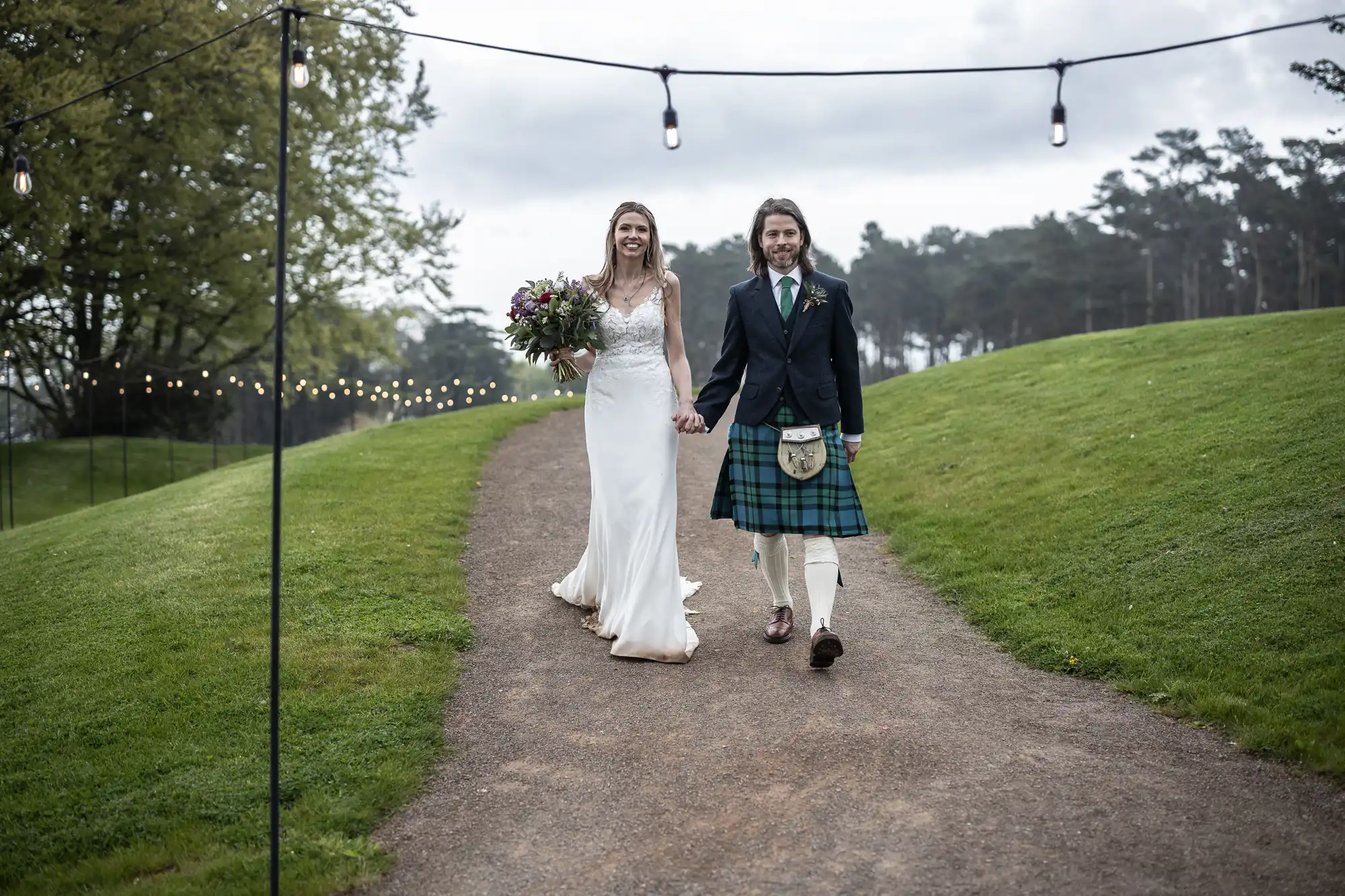 A bride in a white dress and a groom in a kilt walk down an outdoor path lined with string lights, surrounded by green grass and trees.
