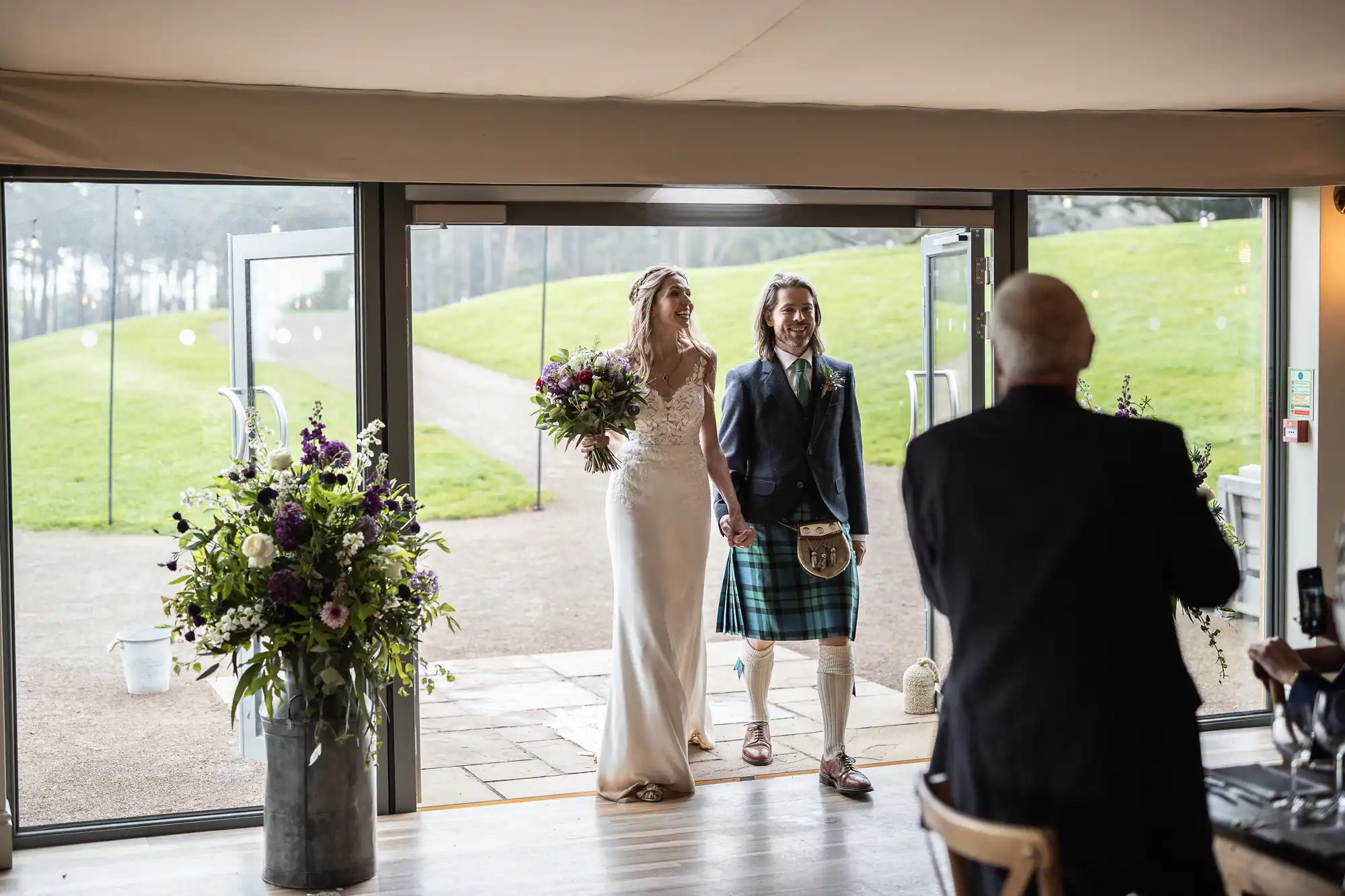 A couple is walking hand-in-hand through a large open door. The woman is wearing a wedding dress and holding a bouquet. The man is dressed in a kilt. An older man is standing nearby with his back to the camera.