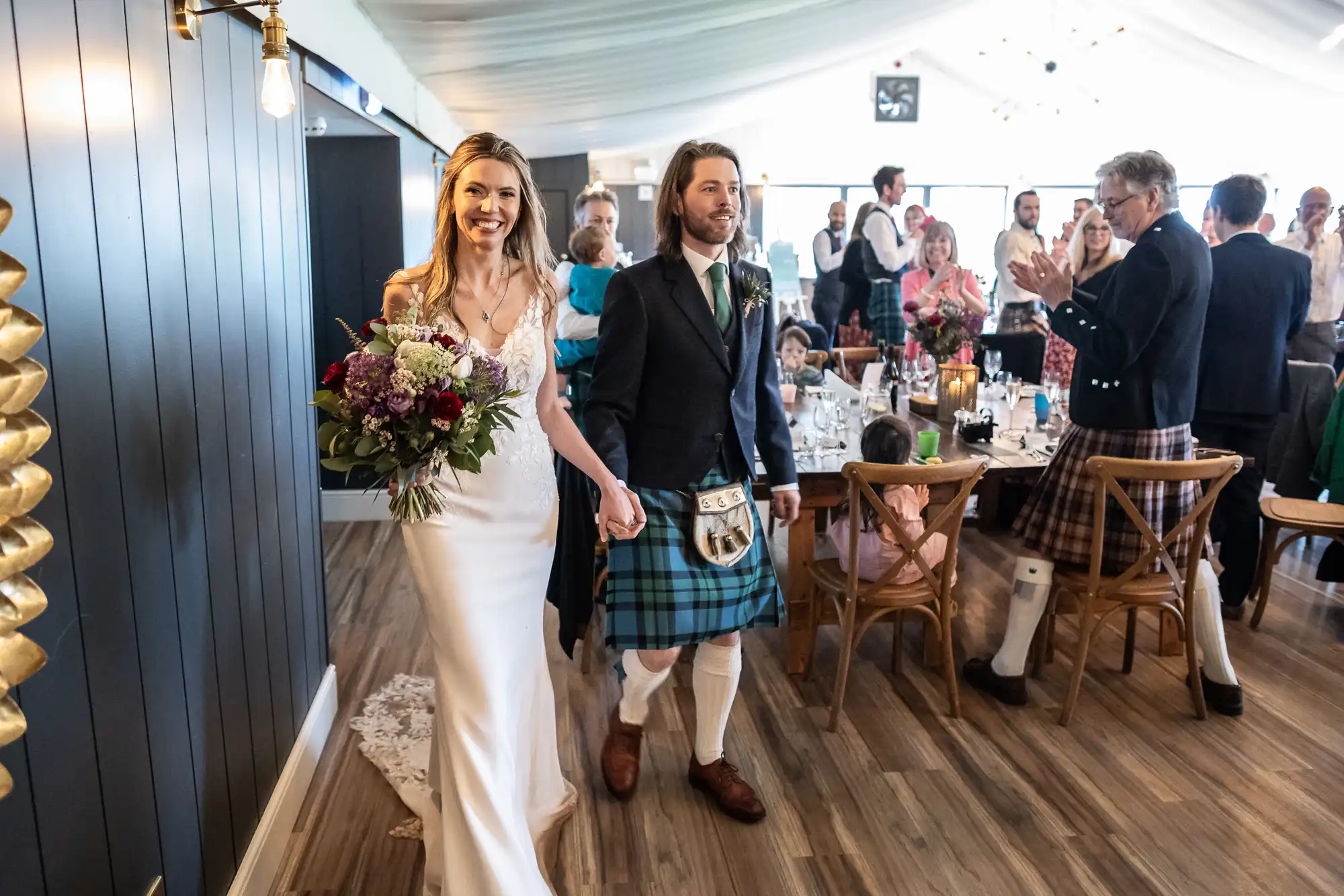 A bride in a white dress and groom in a kilt walk hand in hand in a reception hall, surrounded by applauding guests.