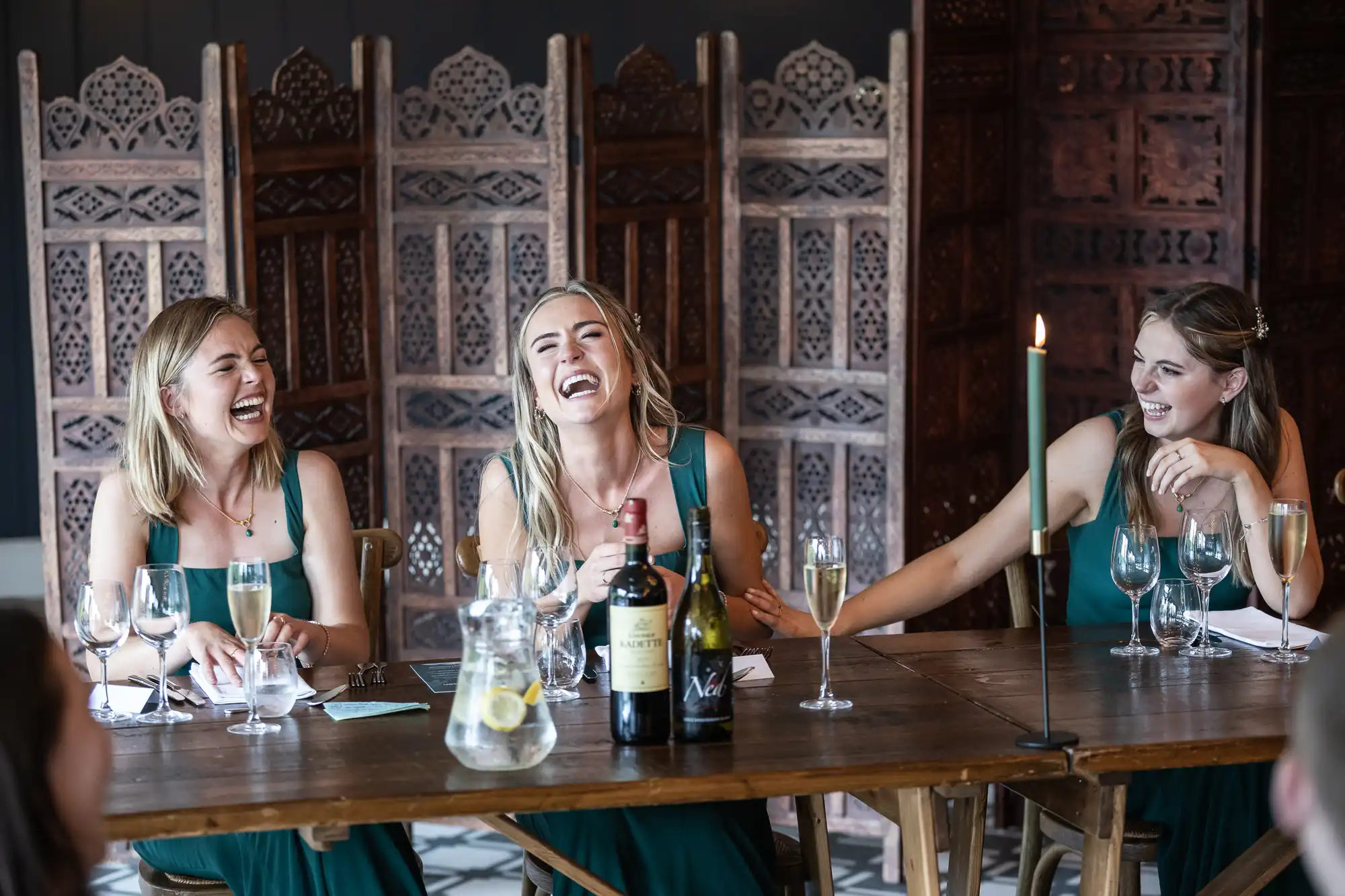 Three women in green dresses are laughing together at a wooden table set with wine, water, and glasses. A candle burns on the table, and decorative wooden panels are in the background.