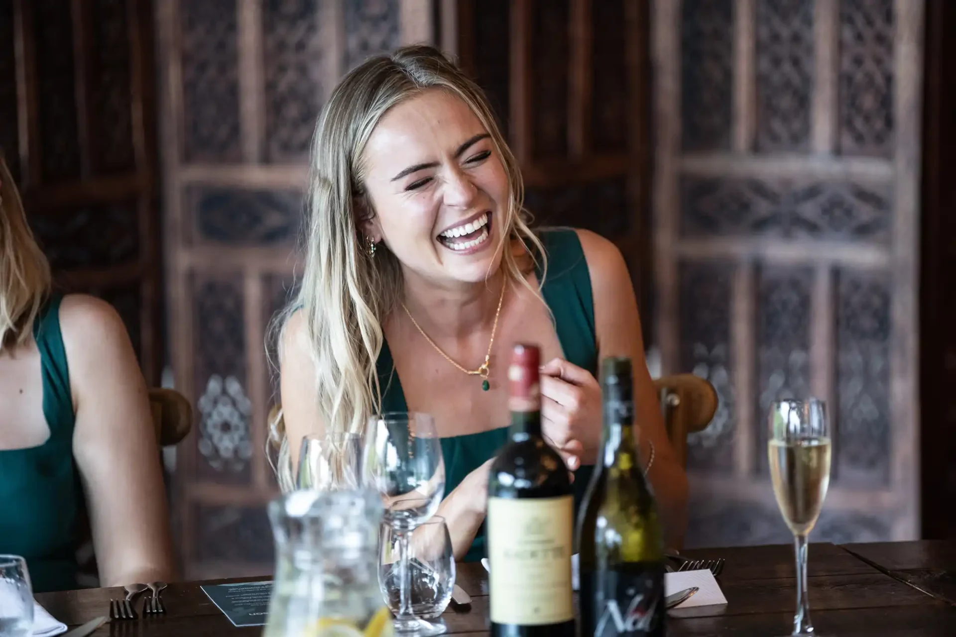 A woman in a green dress sits at a table with wine bottles, a glass of champagne, and a pitcher of water, laughing cheerfully.