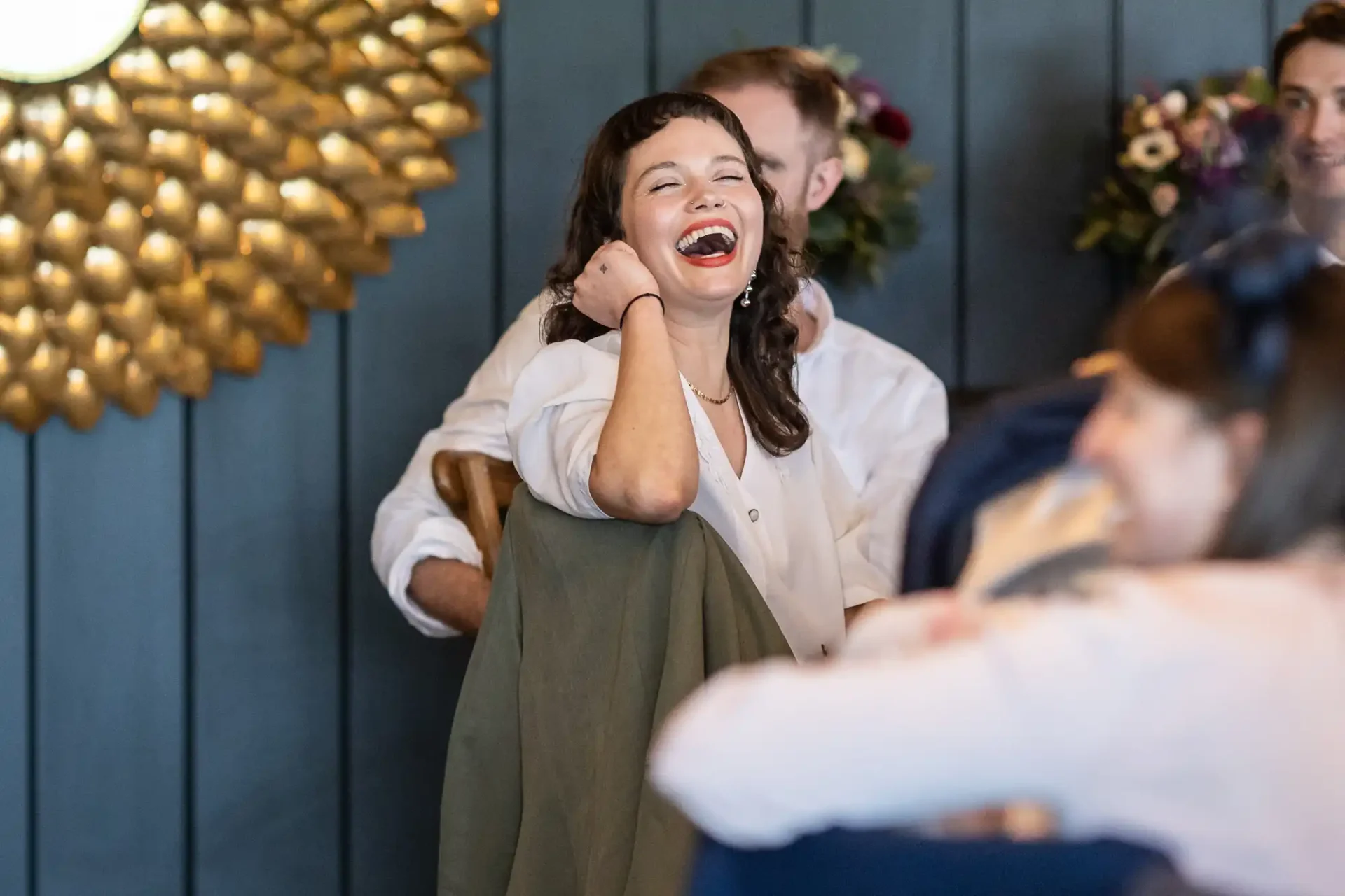 A woman with dark hair laughs heartily while seated among a group of people in a room with dark-paneled walls and floral decor.