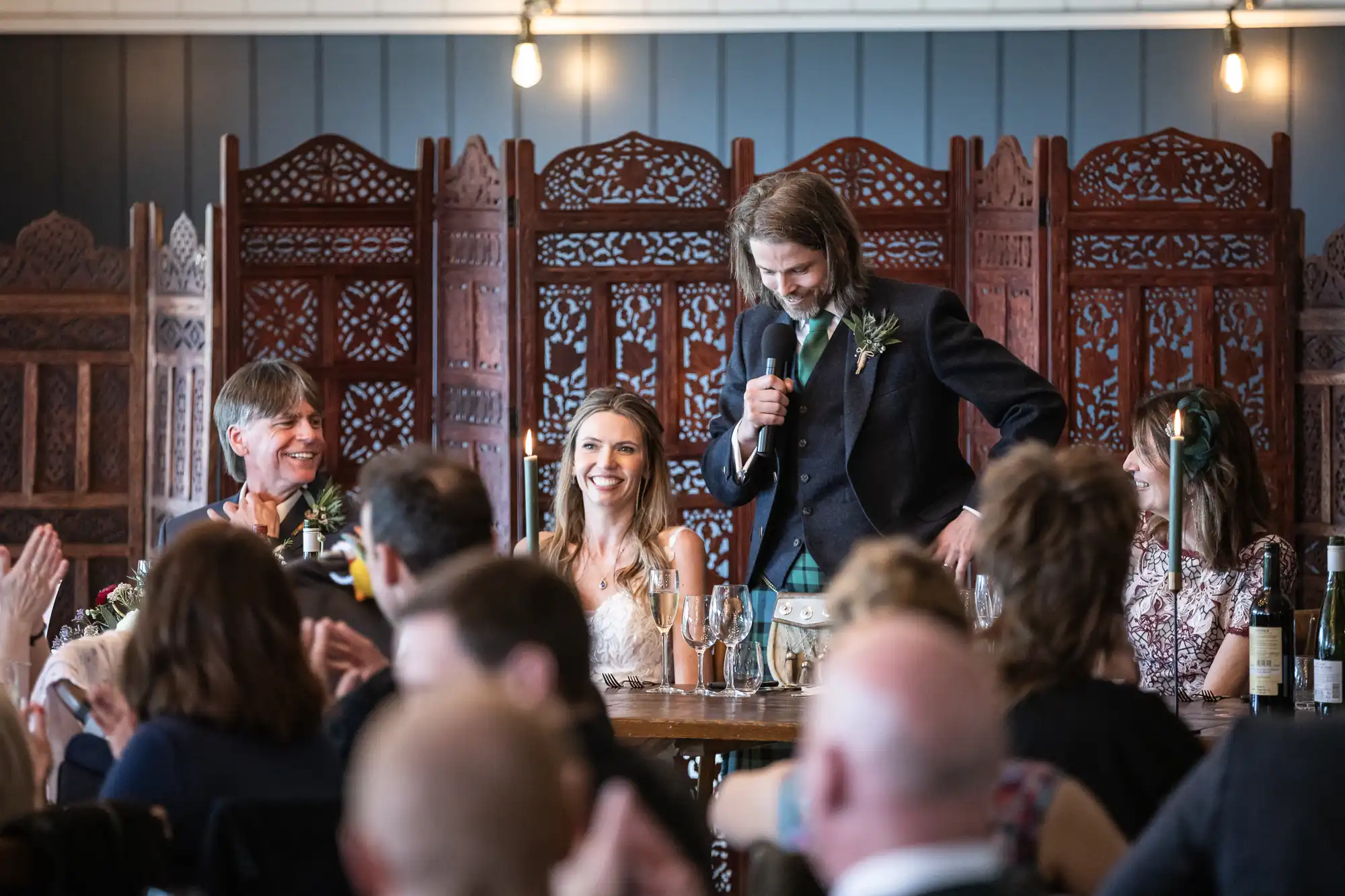A man stands and speaks into a microphone at a formal gathering, while three people sit and smile in front of wooden decorative panels. The room is filled with seated guests.
