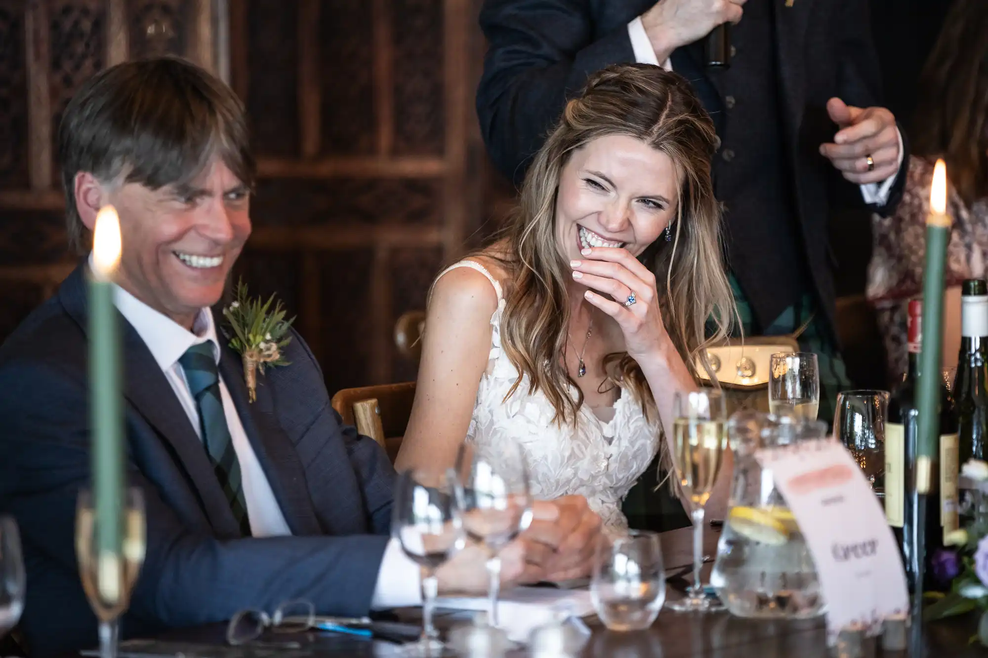A bride and a man in a suit share a laugh at a table set with glasses, bottles, and candles during a wedding reception.