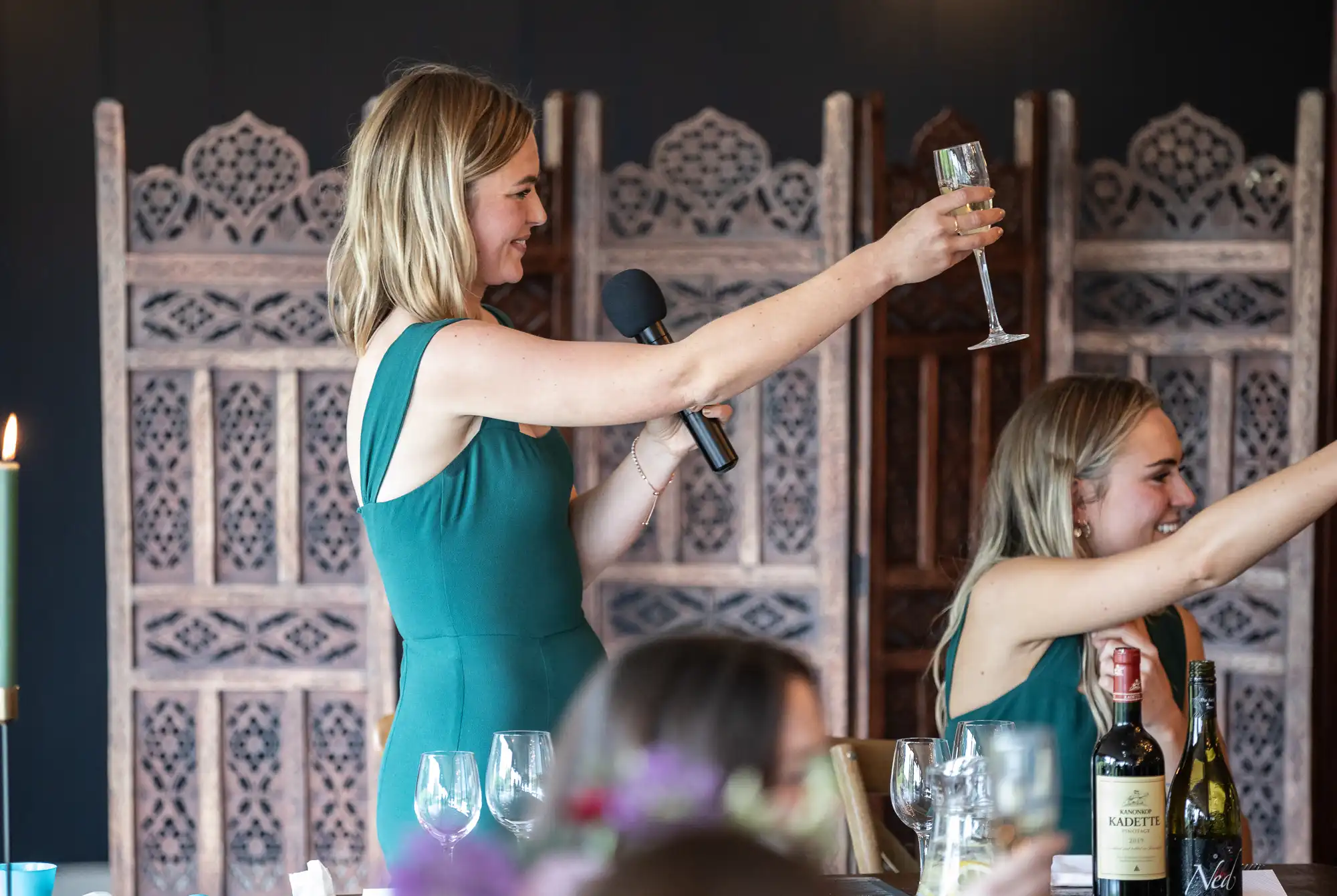 A woman in a green dress holds a microphone and raises a glass for a toast, while another woman in a similar dress also raises her glass at a dining table with bottles and glasses.