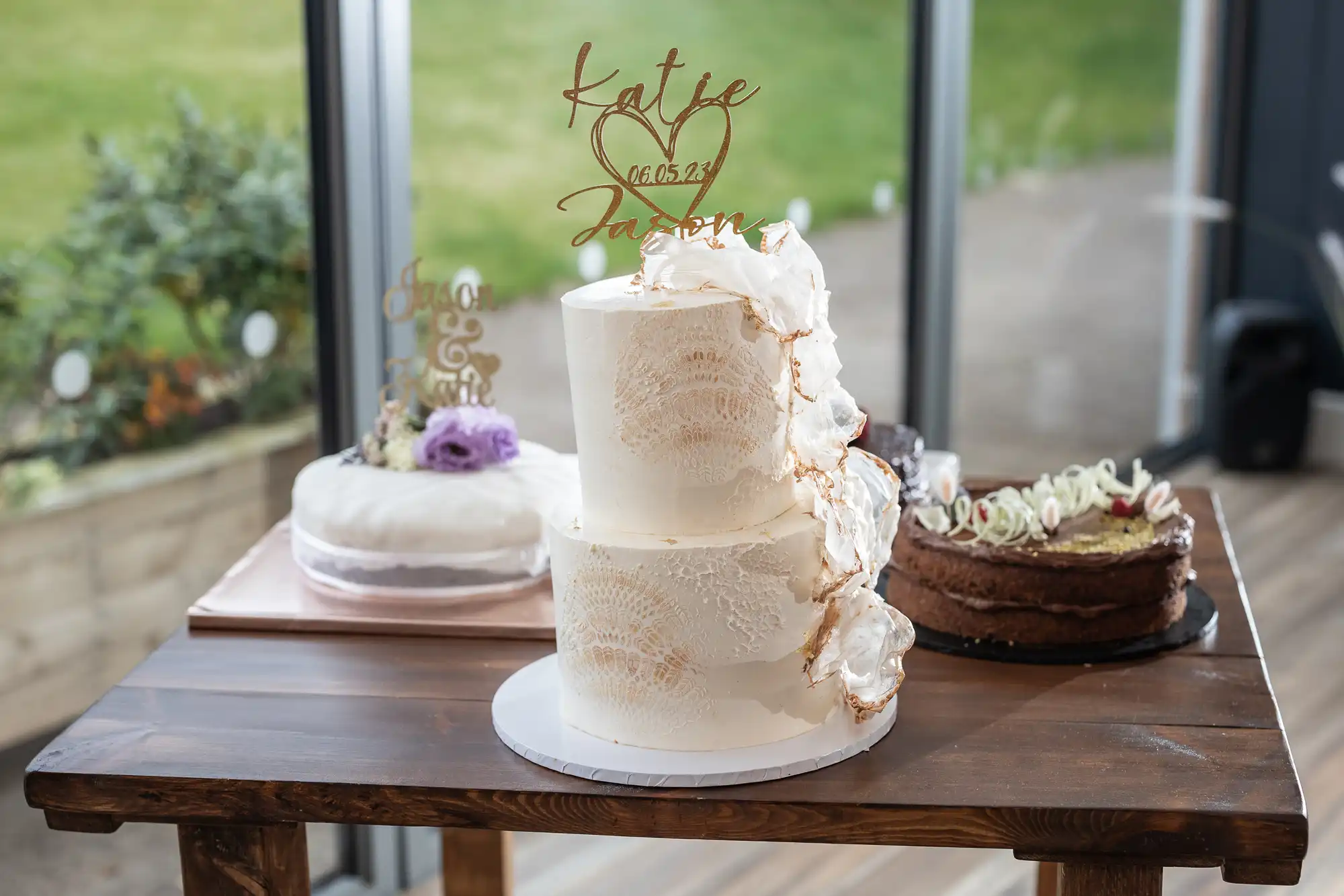An elegantly decorated wedding cake with "Katie & Jason, 16-05-23" topper. Additional cakes are on the same table. The background features a garden seen through windows.