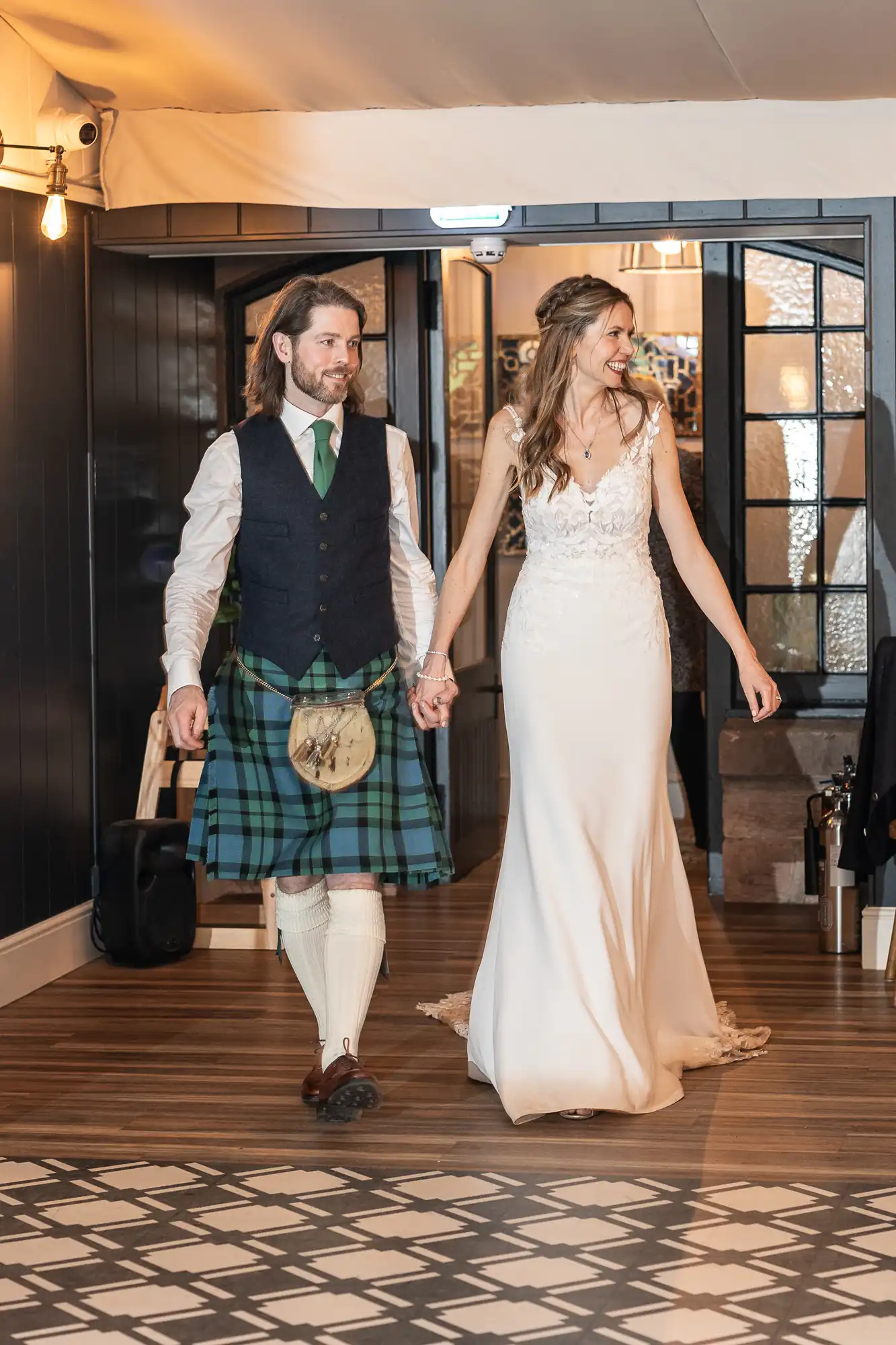 A couple, with the man in a kilt and the woman in a white dress, walk hand in hand through a doorway at an indoor event.