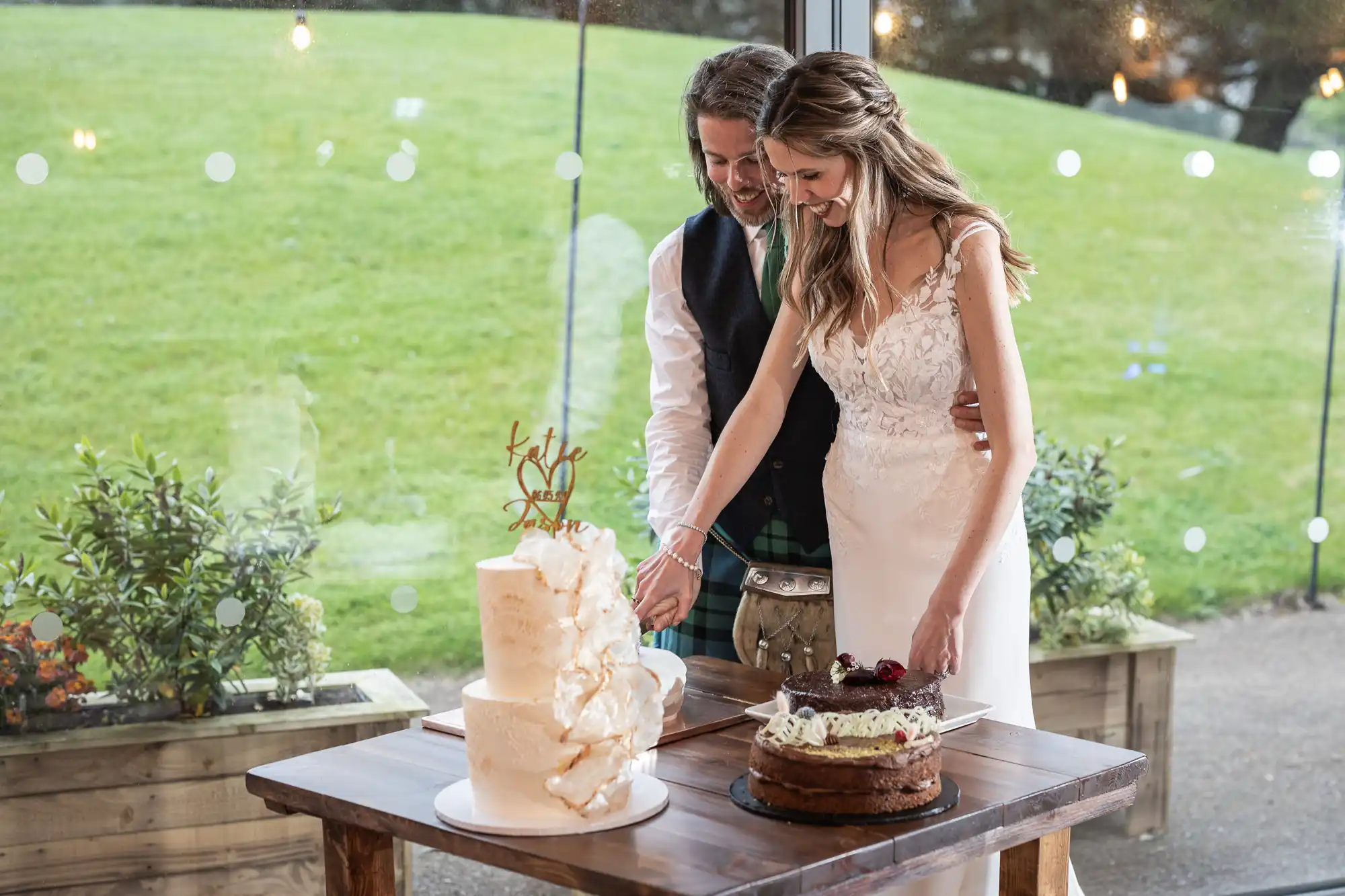 A couple cuts their wedding cake together. They stand in front of a window with a grassy hill outside. There are two cakes on the table; one is tiered and white, the other is a single-layer chocolate cake.
