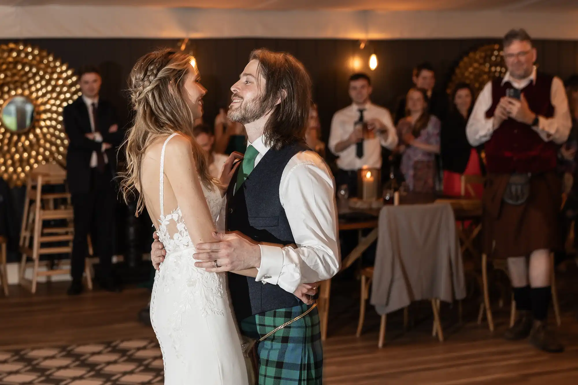 A bride and groom dance together, with guests watching in the background. The groom is wearing a kilt, and the event appears to be a wedding reception.